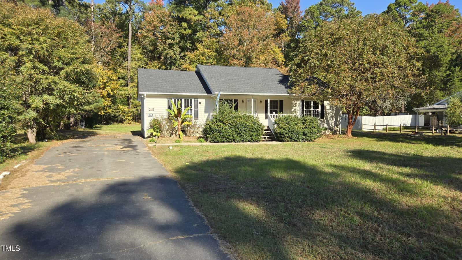A charming single-story house at 1408 W B Street in Butner features a gray roof and white siding, nestled among grass and trees. A paved driveway leads to the entrance, flanked by a few shrubs. In the background, additional trees display vibrant autumn foliage.