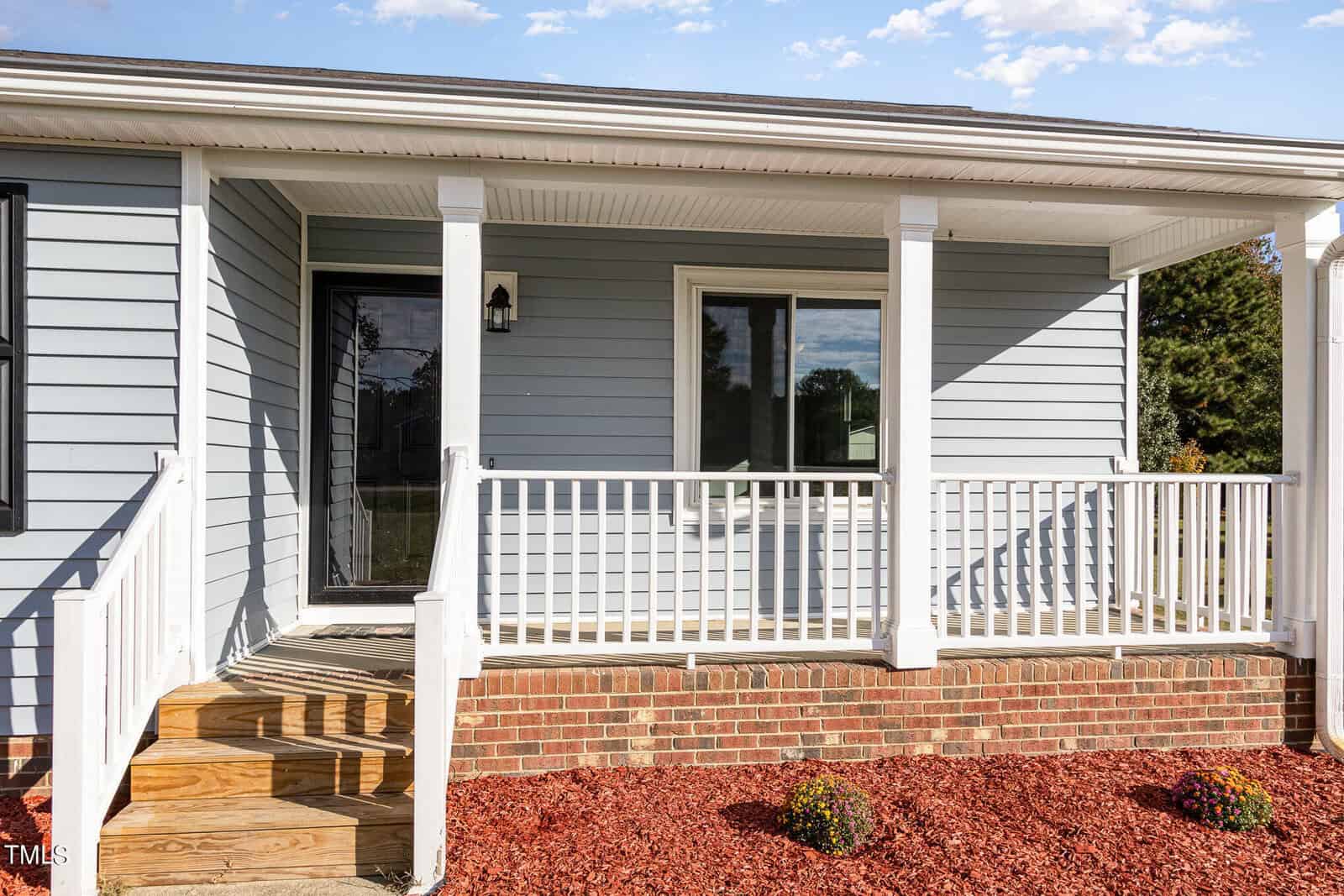 A small front porch with white railing, steps leading to a black front door, a window to the right, light gray siding, brick foundation, and mulch with small plants in front.