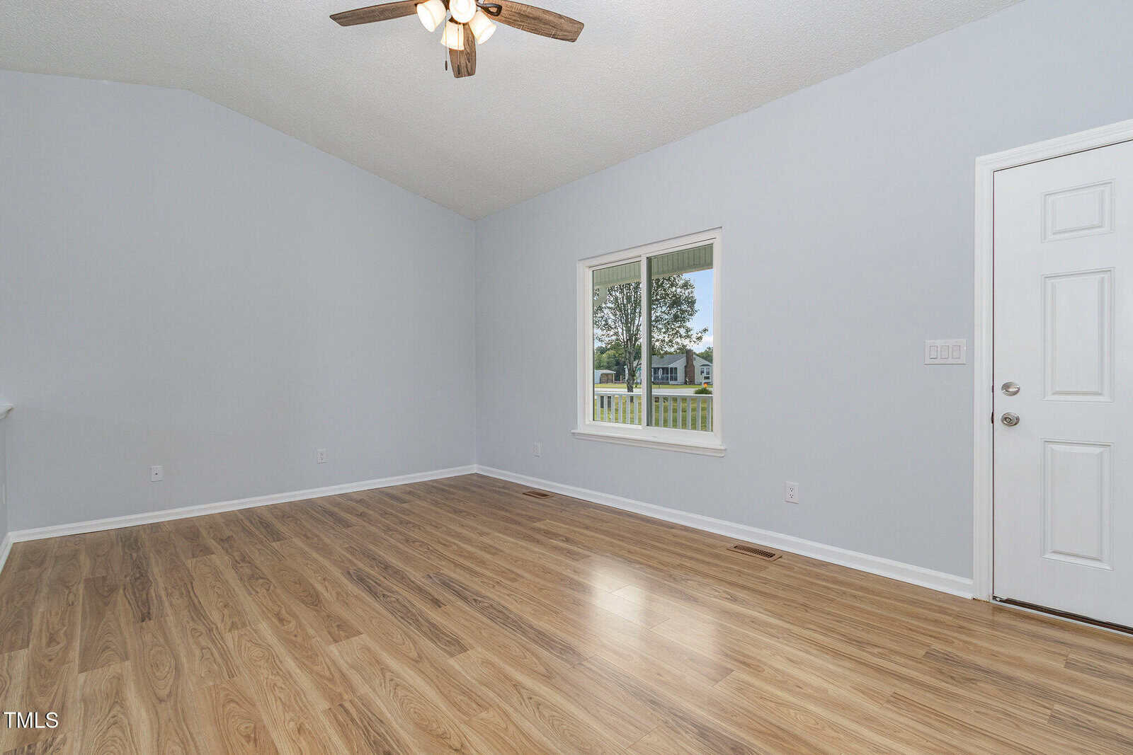 A spacious empty room with light gray walls, wood-look flooring, a ceiling fan with lights, a white-framed window, and a white door. Outside the window, a green lawn and trees are visible.