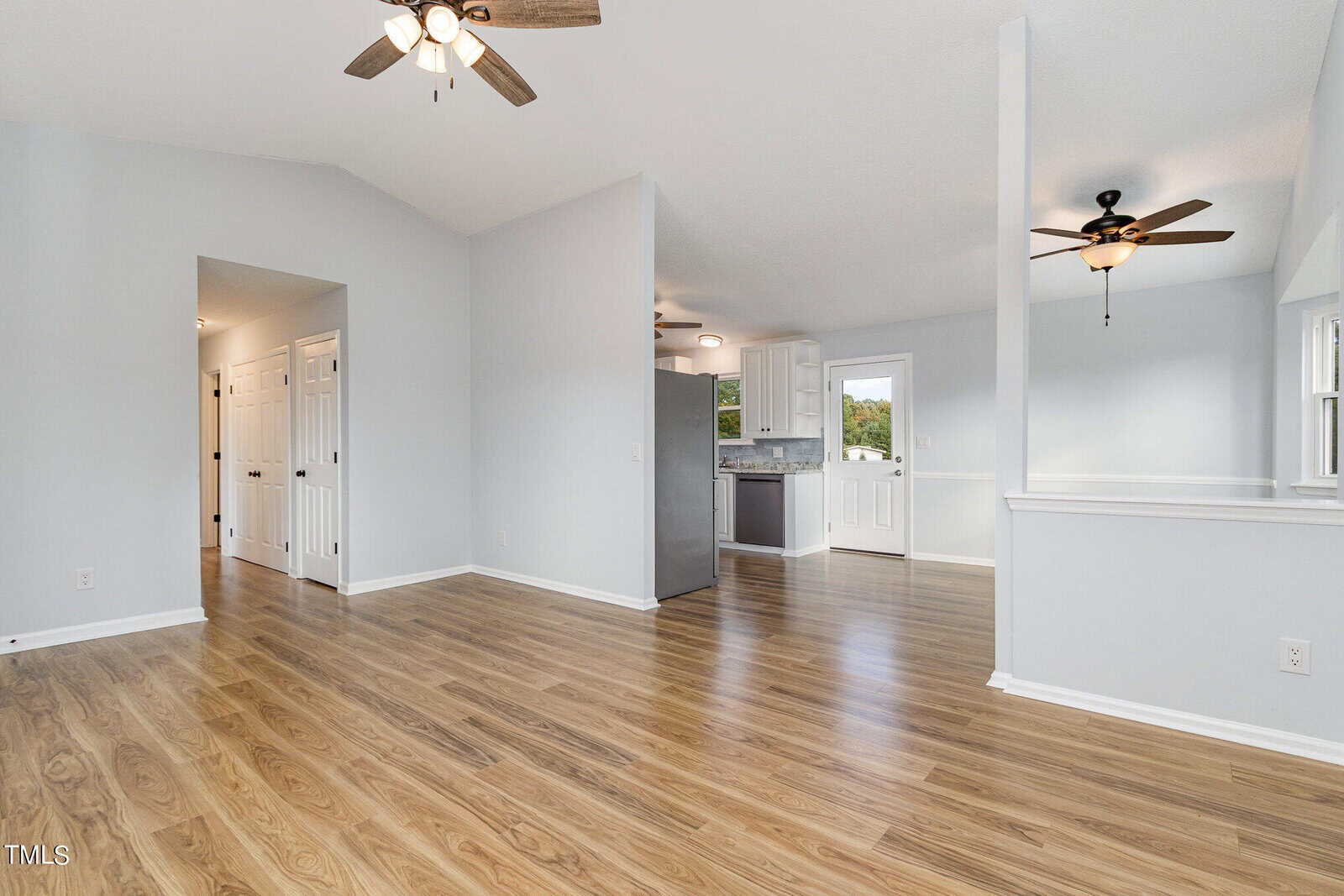 Spacious room with light wood flooring, white walls, and two ceiling fans. Kitchen with white cabinets and stainless steel appliances is visible. Windows and doors provide natural light.