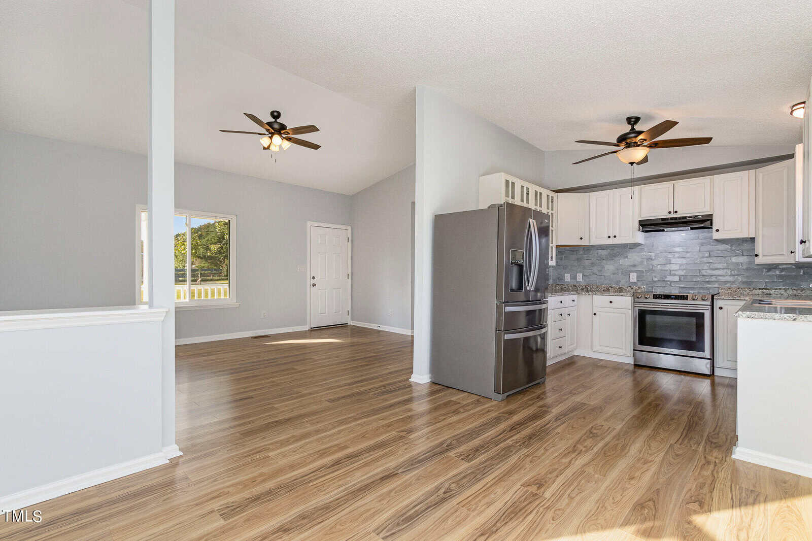 Open-concept kitchen and living area with light-colored wood flooring, stainless steel appliances, white cabinets, gray backsplash, ceiling fans, and a window and door in the background.