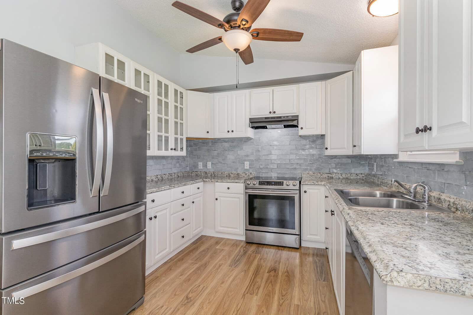 Modern kitchen with stainless steel refrigerator, oven, and range hood, white cabinets, glass-front upper cabinets, granite countertops, light gray tile backsplash, ceiling fan, and wood flooring.