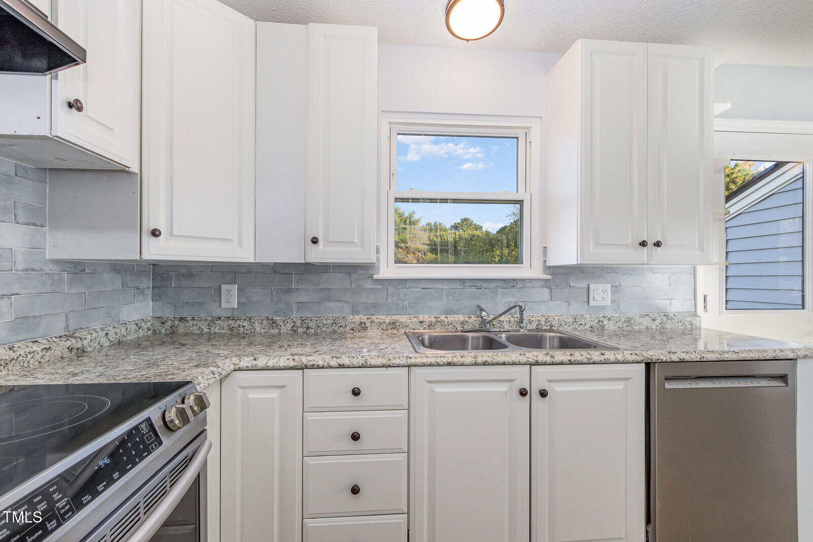 A modern kitchen with white cabinets, granite countertops, a double sink under a window, stainless steel dishwasher, and a stove. The backsplash is gray tile, and natural light enters through the window.
