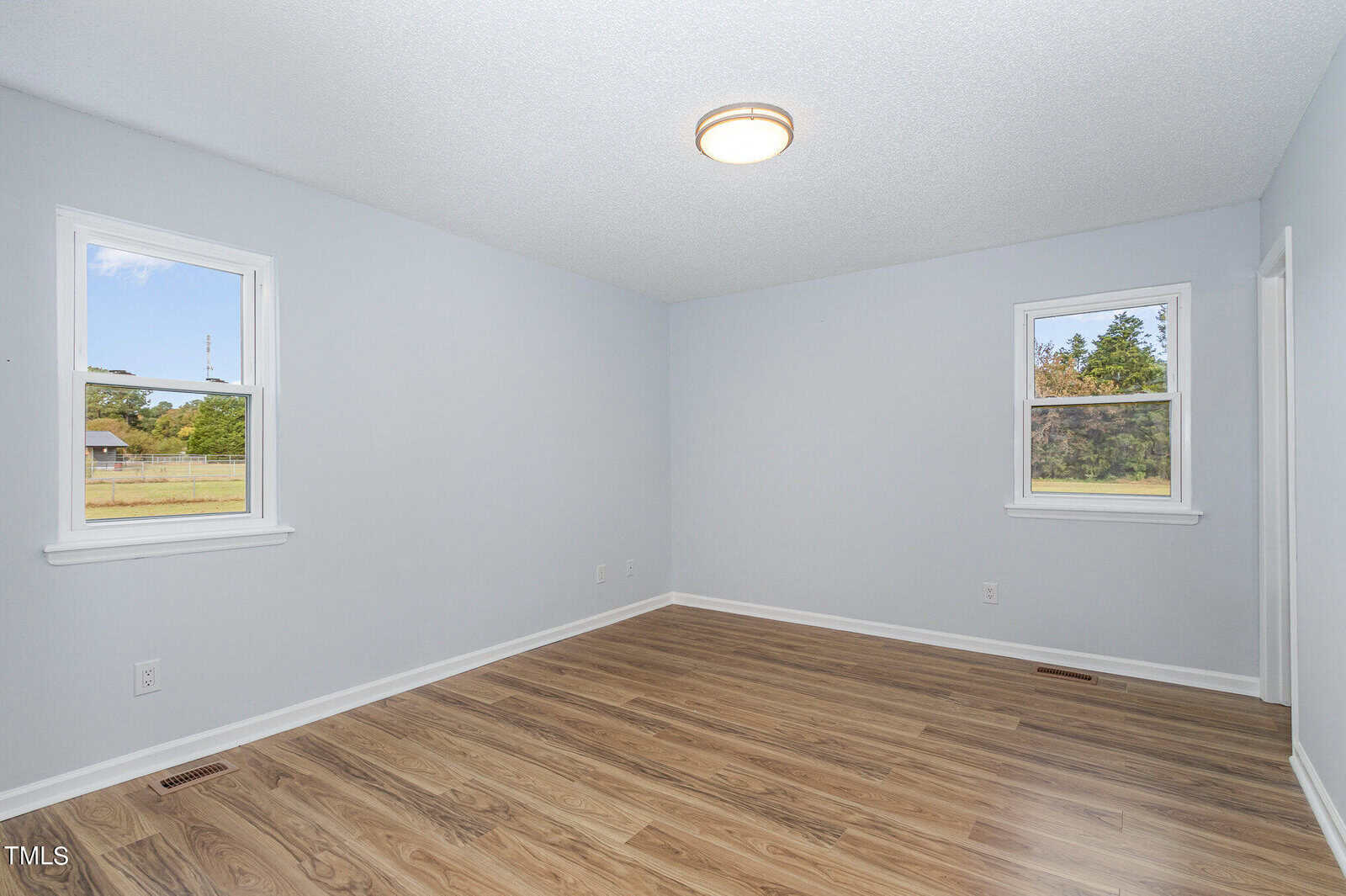 Empty room with light gray walls, wood-style flooring, two windows with outdoor views, a white ceiling with a round light fixture, and no furniture or decor.