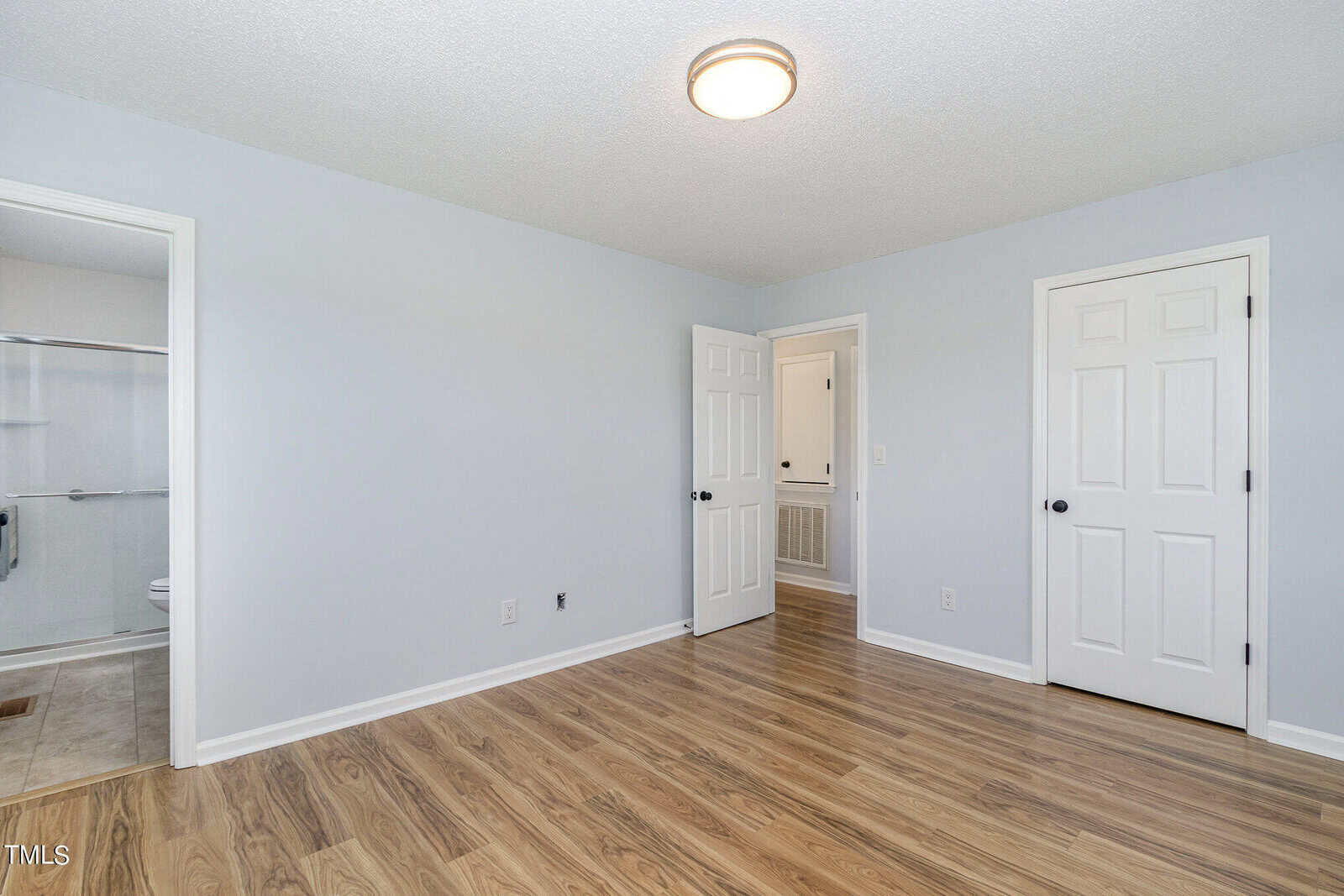 Empty bedroom with light gray walls, wood flooring, white trim, a ceiling light, two closed white doors, and an open door leading to a bathroom with a glass shower.