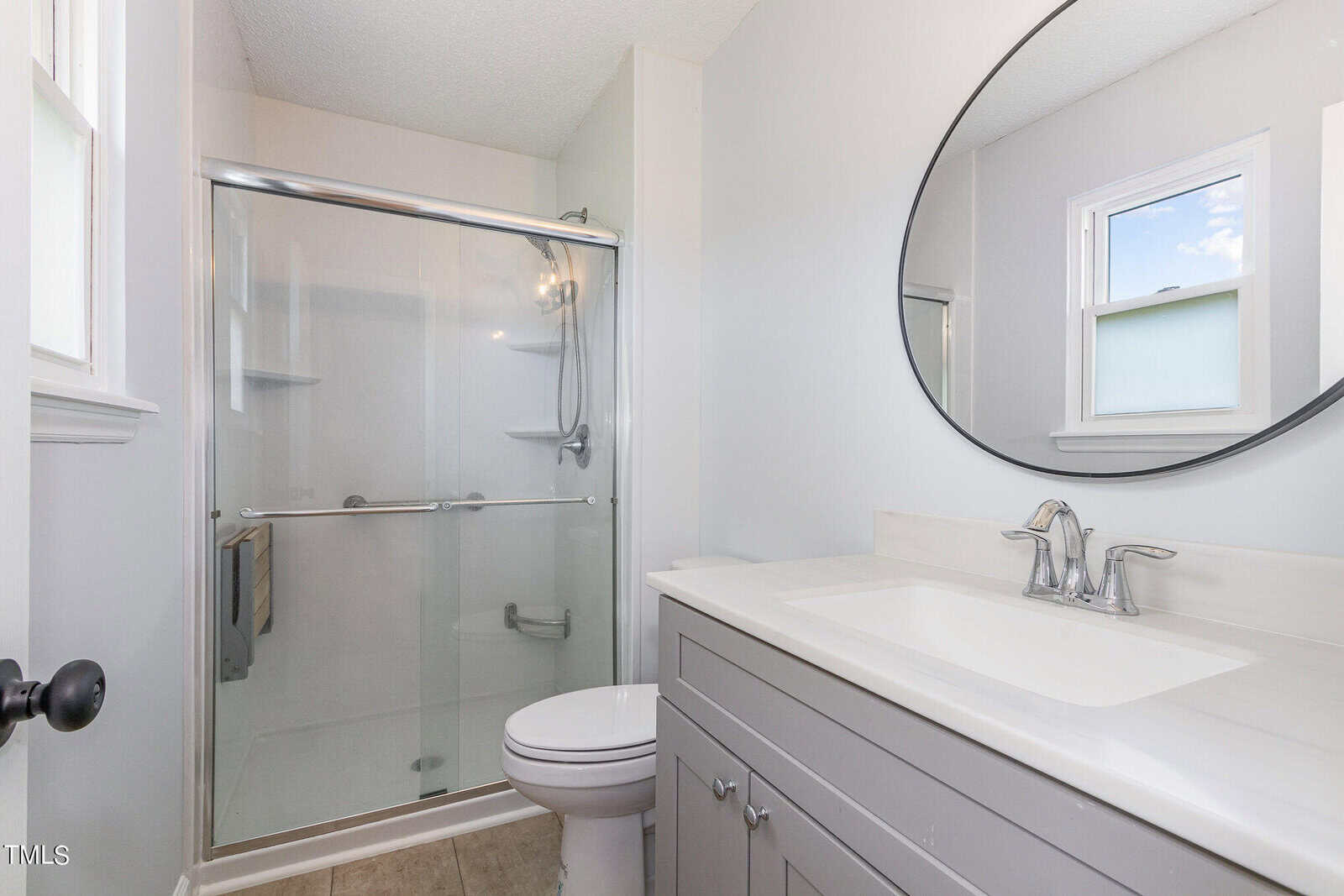 Modern bathroom with a glass-enclosed shower, gray vanity with sink, silver fixtures, oval mirror, and two windows letting in natural light.