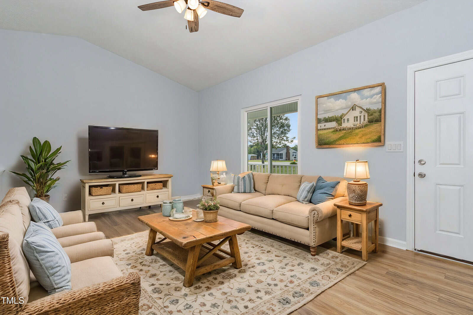 A living room with light blue walls, two beige sofas, a wooden coffee table, a TV on a wooden stand, a ceiling fan, two side tables with lamps, a plant, a window, a door, and a framed painting on the wall.