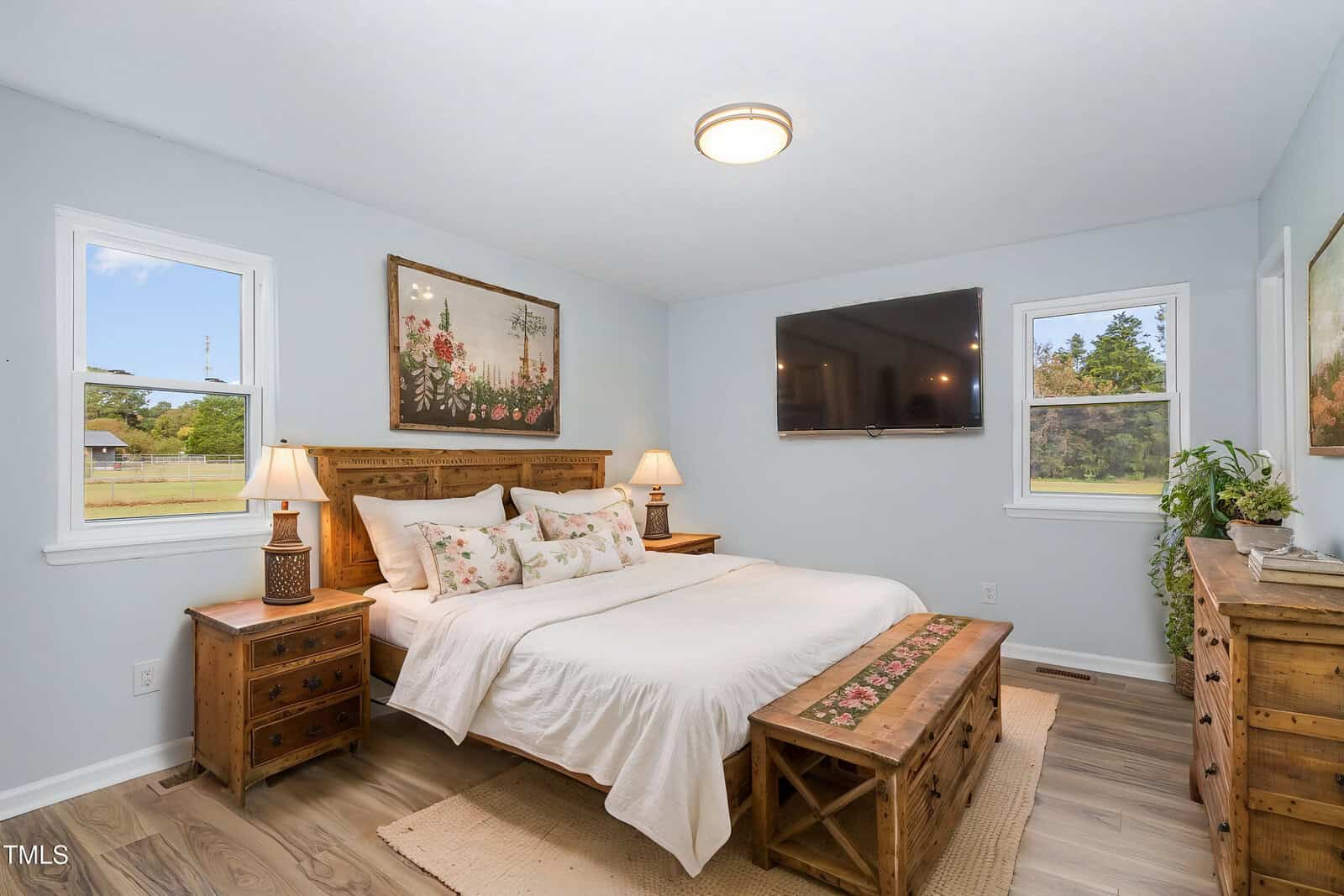 A bedroom with a wooden bed frame, two matching nightstands, a bench at the foot of the bed, a large wall-mounted TV, two windows, and a floral painting above the headboard. The room has light-colored walls and wood flooring.