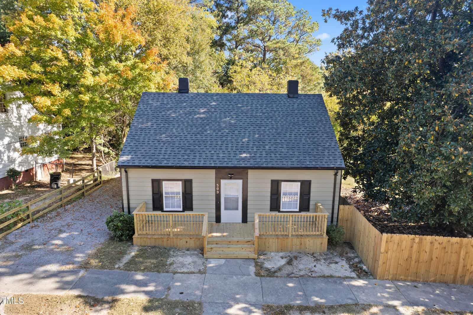 A small beige house with a dark gray roof and two front windows, surrounded by a wooden fence. A pathway leads to the front door, and there are trees with autumn foliage in the background.