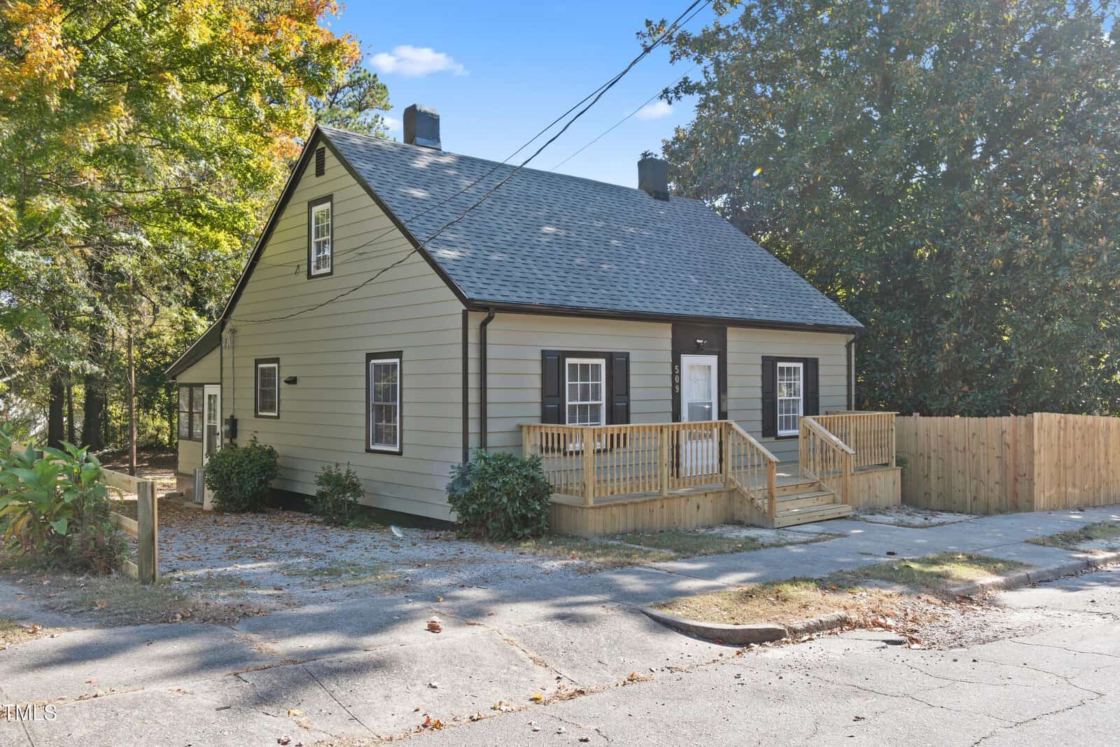 A beige single-story house with a dark gray roof and white-framed windows. It features a small porch with steps leading up to the front door. The house is surrounded by trees and a wooden fence. The street in front is lined with fallen leaves.