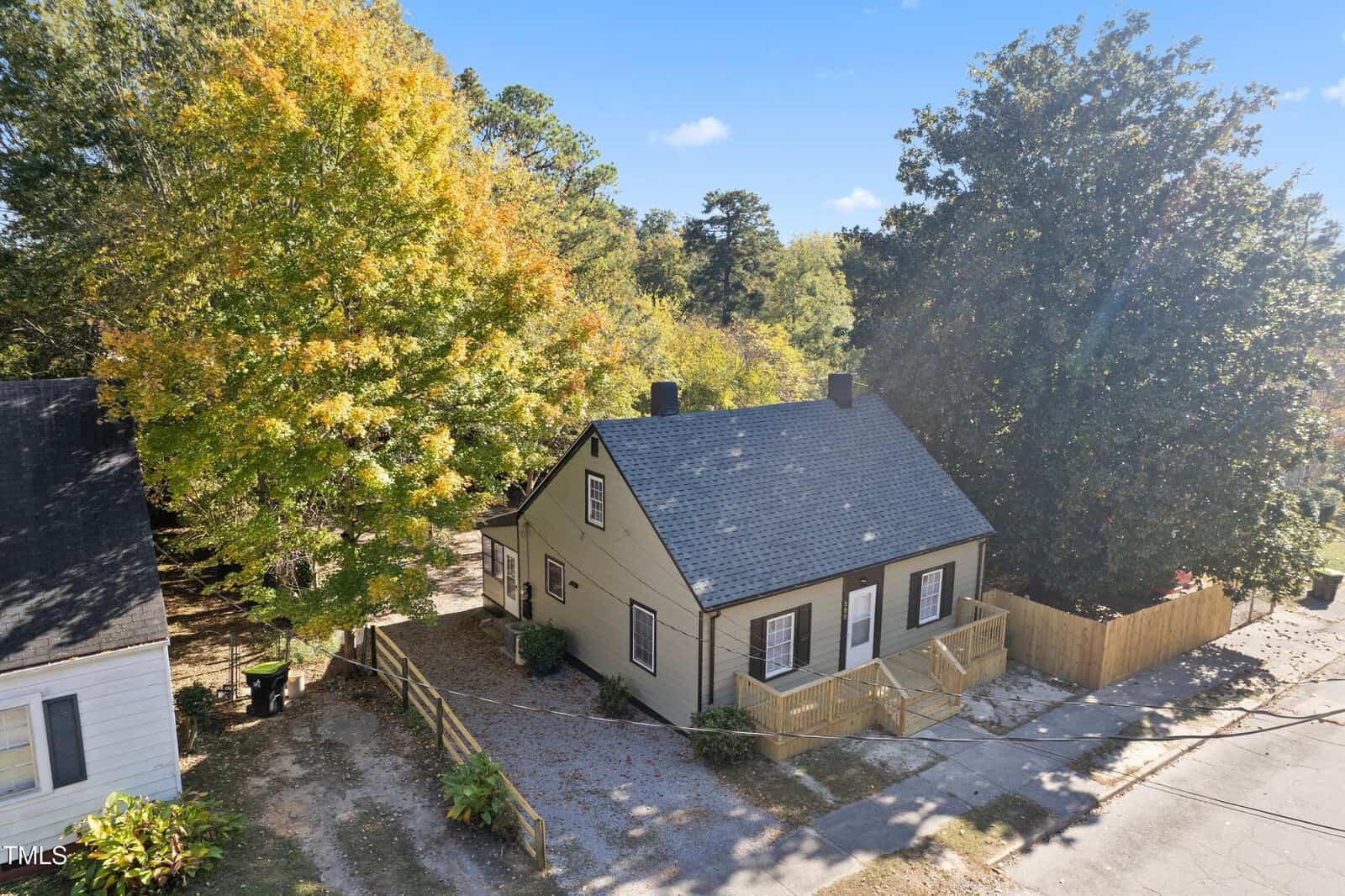 Aerial view of a small, beige house with a gray roof, surrounded by trees with autumn foliage. The house has a simple wooden fence and a driveway. Neighboring homes are partially visible, and the sky is clear with a few clouds.