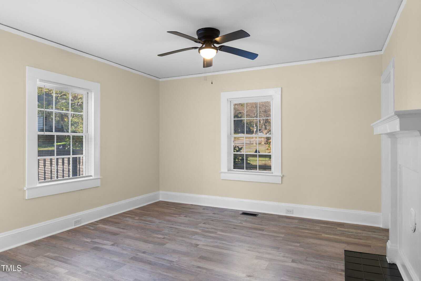 An empty living room with light-colored walls and wood flooring. The room features large windows with white trim, allowing natural light to enter. A ceiling fan with a light is centered on the ceiling. There is a white fireplace on the right.