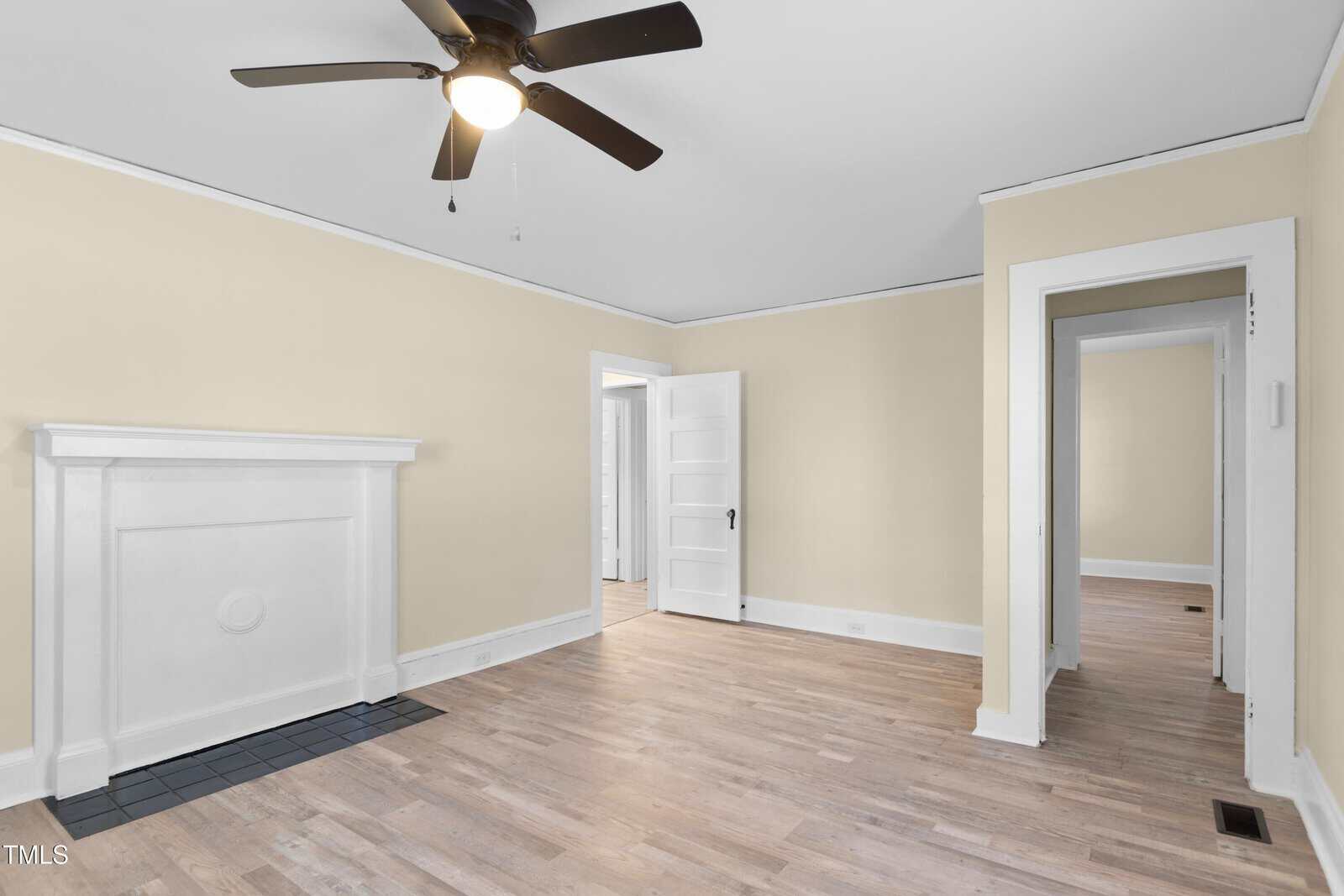 An empty room with pale yellow walls, a white ceiling, and light wood flooring. A white fireplace is on the left wall. A black ceiling fan hangs from the ceiling. Two doorways are visible, one leading to another room.