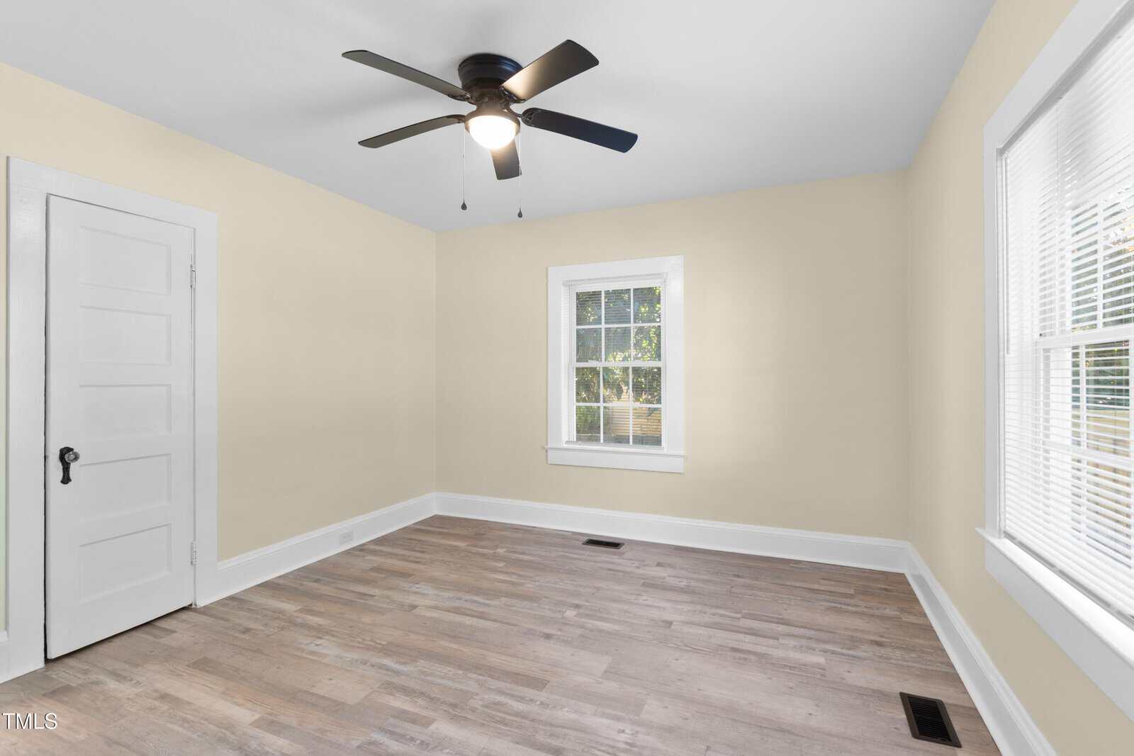 A bright, empty room with light yellow walls and wooden flooring. A window with white blinds is on the right wall, and a white door is on the left. A ceiling fan with a light is centered on the ceiling.