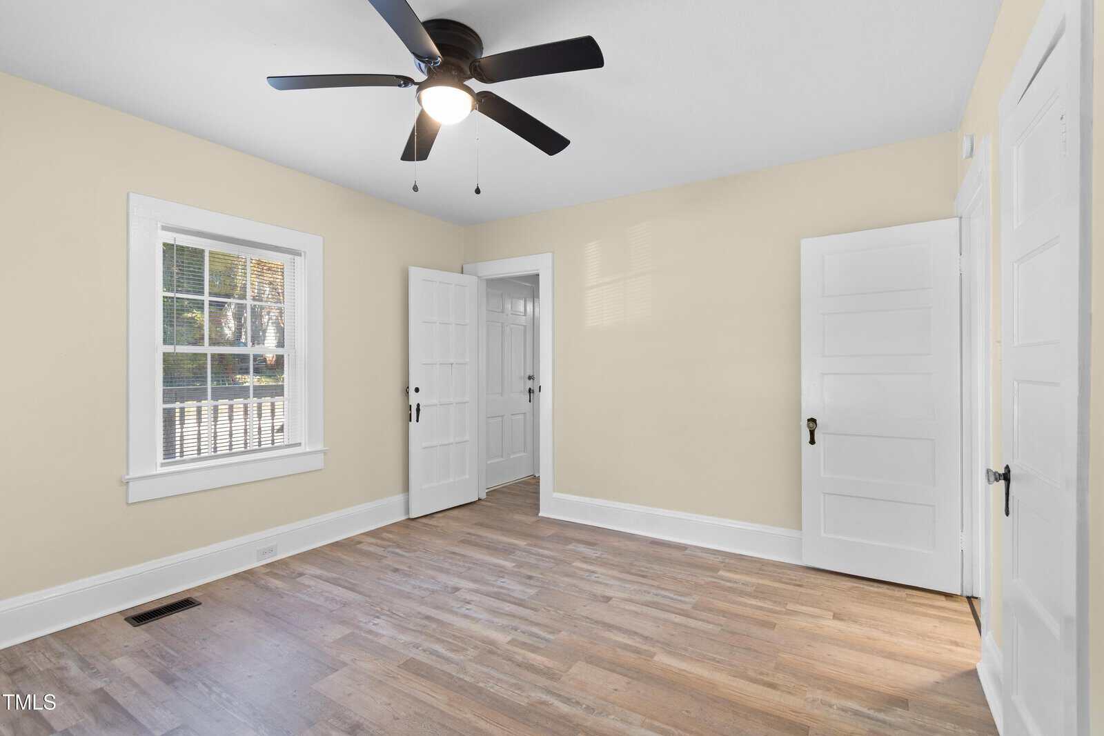 A bright, empty room with light yellow walls, a white ceiling, and wood laminate flooring. There's a ceiling fan with black blades, two white doors, and a window with white blinds. Natural light streams in through the window.