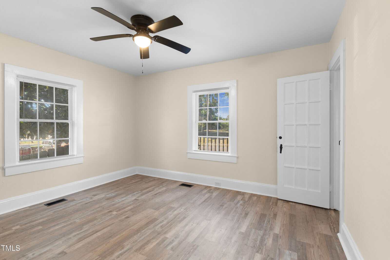 A bright, empty room features light beige walls and two large windows with white trim, allowing natural light to stream in. The hardwood floor adds warmth, and a ceiling fan hangs from the white ceiling. A white door is on the right side of the room.