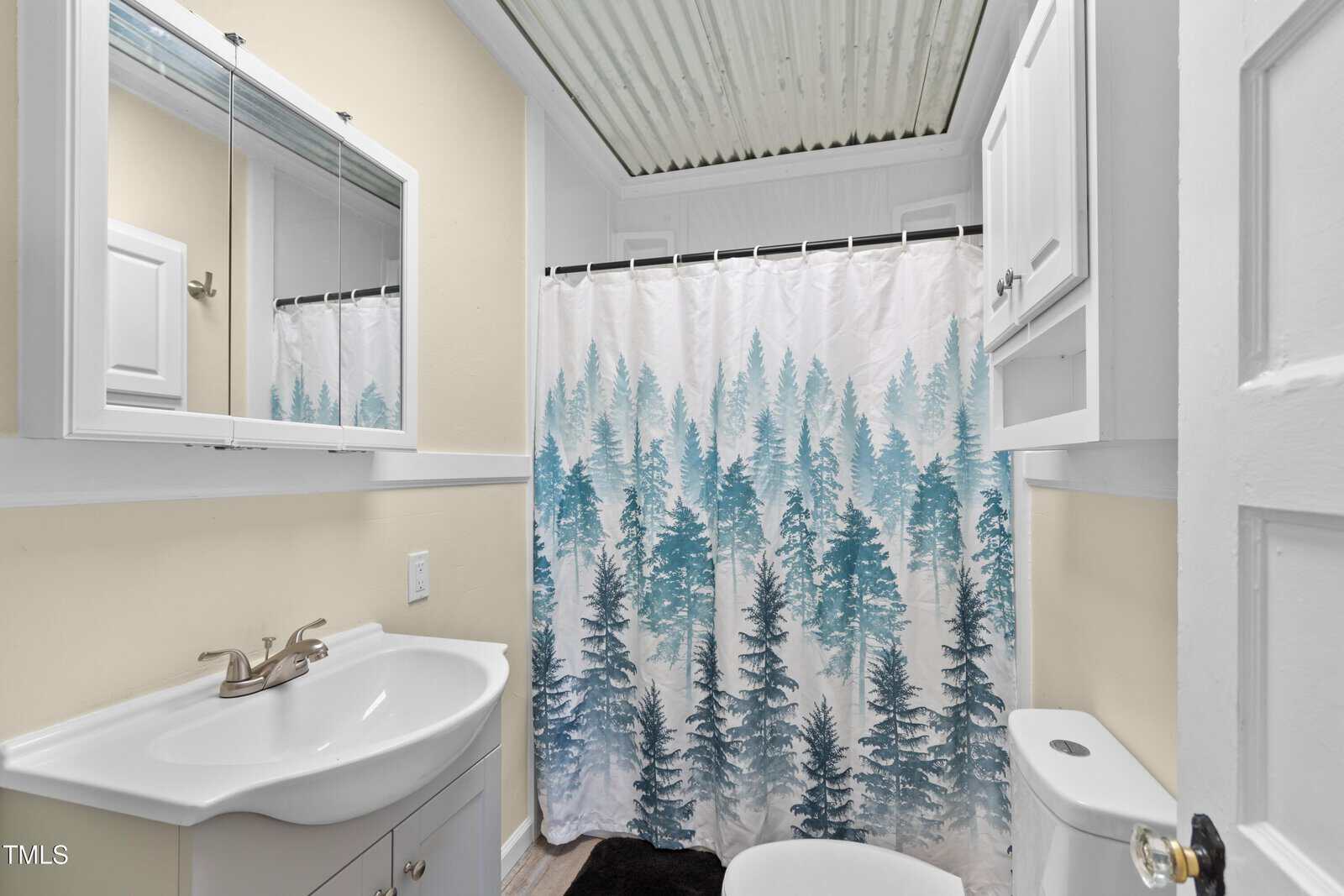 A small bathroom with a white sink and cabinet, decorative shower curtain featuring blue and green trees, and a white toilet. The walls are light yellow, and the ceiling has a corrugated pattern.