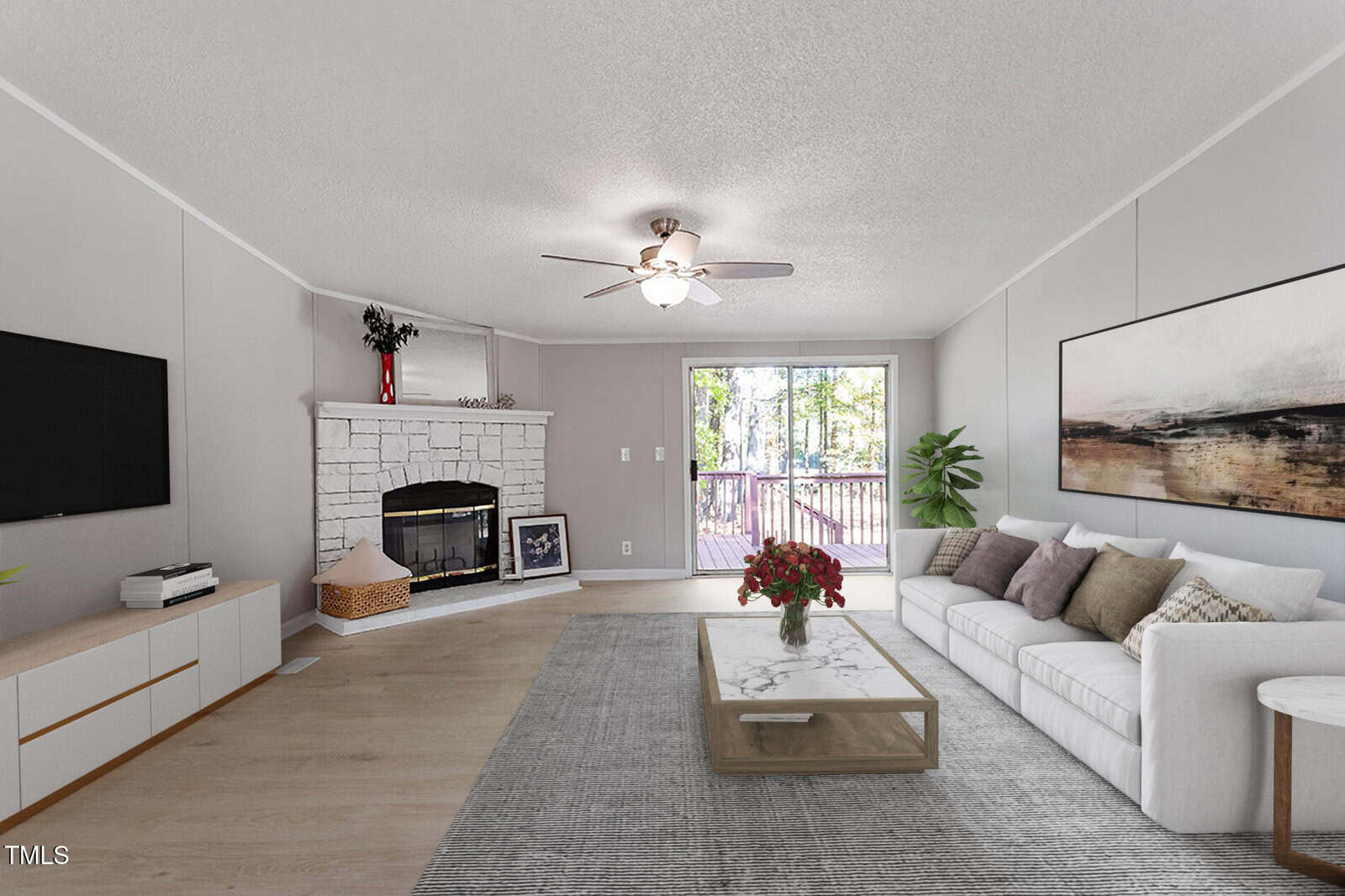 A living room with a white couch, a coffee table with flowers, a gray rug, a wall-mounted TV, a stone fireplace, large artwork, and glass doors leading to a deck with trees visible outside.