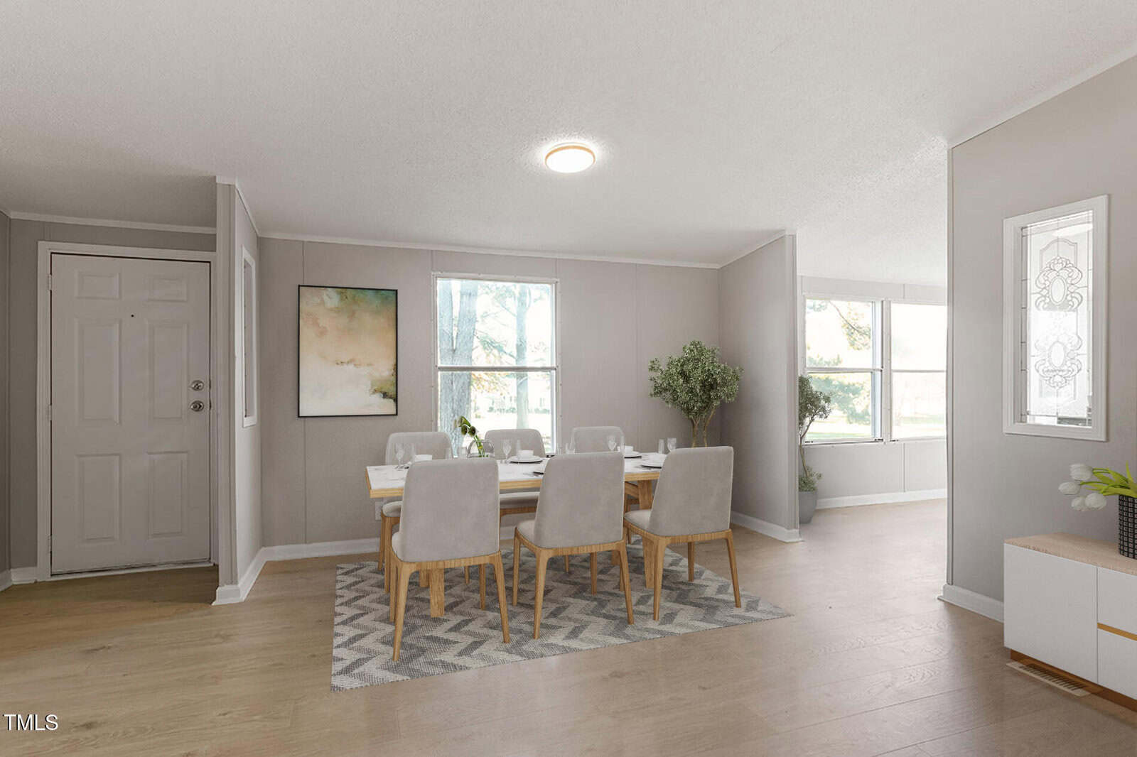 A dining room with a rectangular table set for six, light wood chairs with fabric seats, a modern rug, and wall art. Large windows let in natural light. The space has light walls and wood floors. A door is visible to the left.