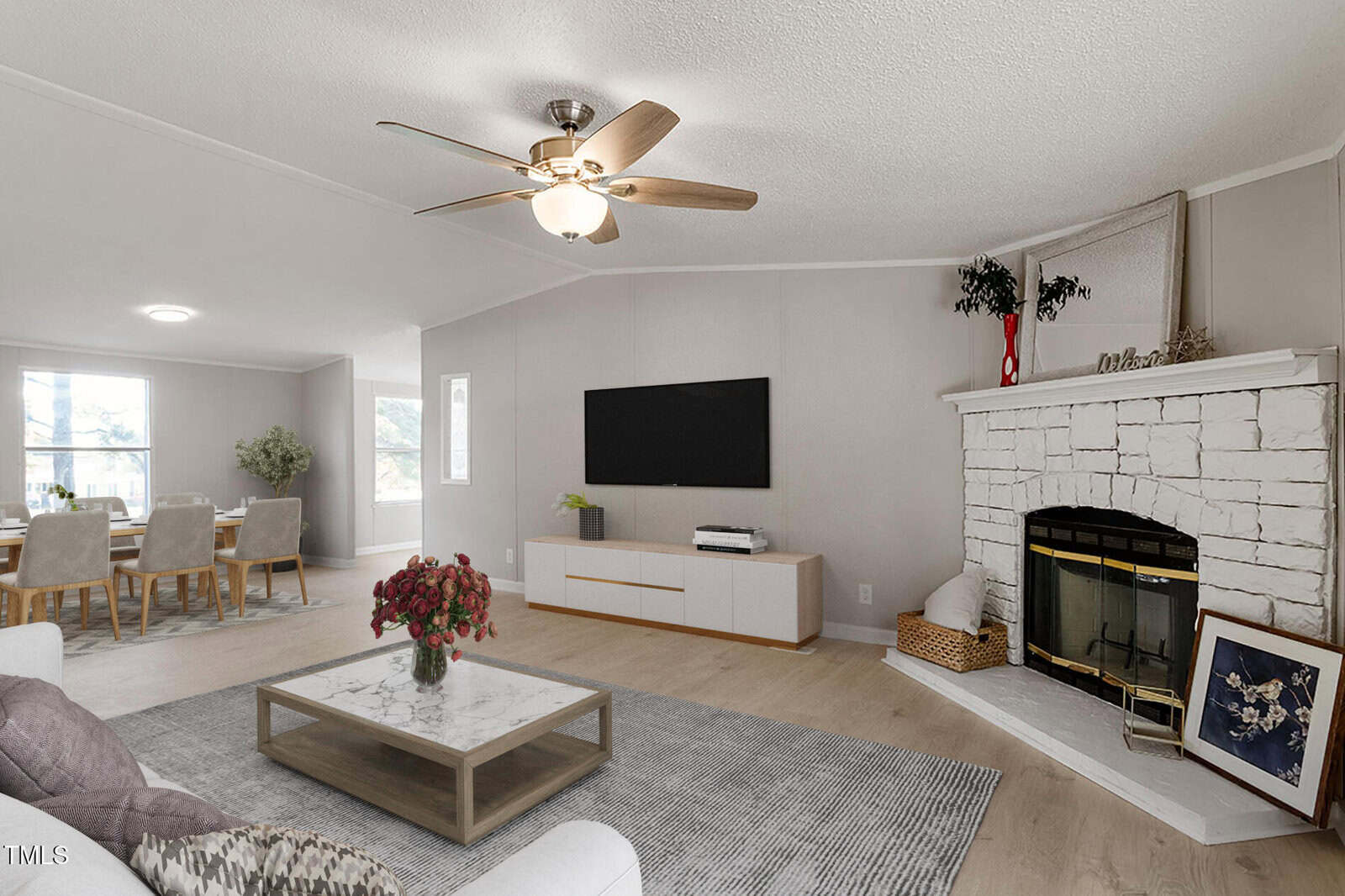 A modern living room with light wood floors, a white stone fireplace, a wall-mounted TV, a coffee table, and a dining area in the background. The space is decorated with minimalist furniture and neutral tones.