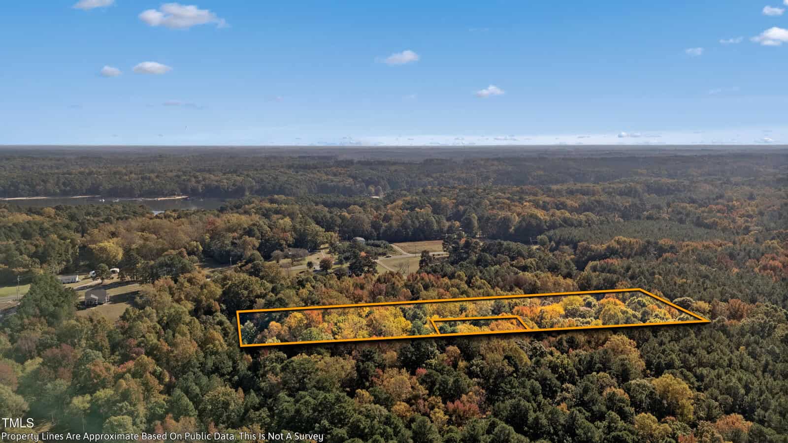 Aerial view of a wooded landscape during autumn, with a rectangular area outlined in orange. The sky is clear with scattered clouds. The foliage displays a range of colors from green to brown. A body of water is visible in the background.
