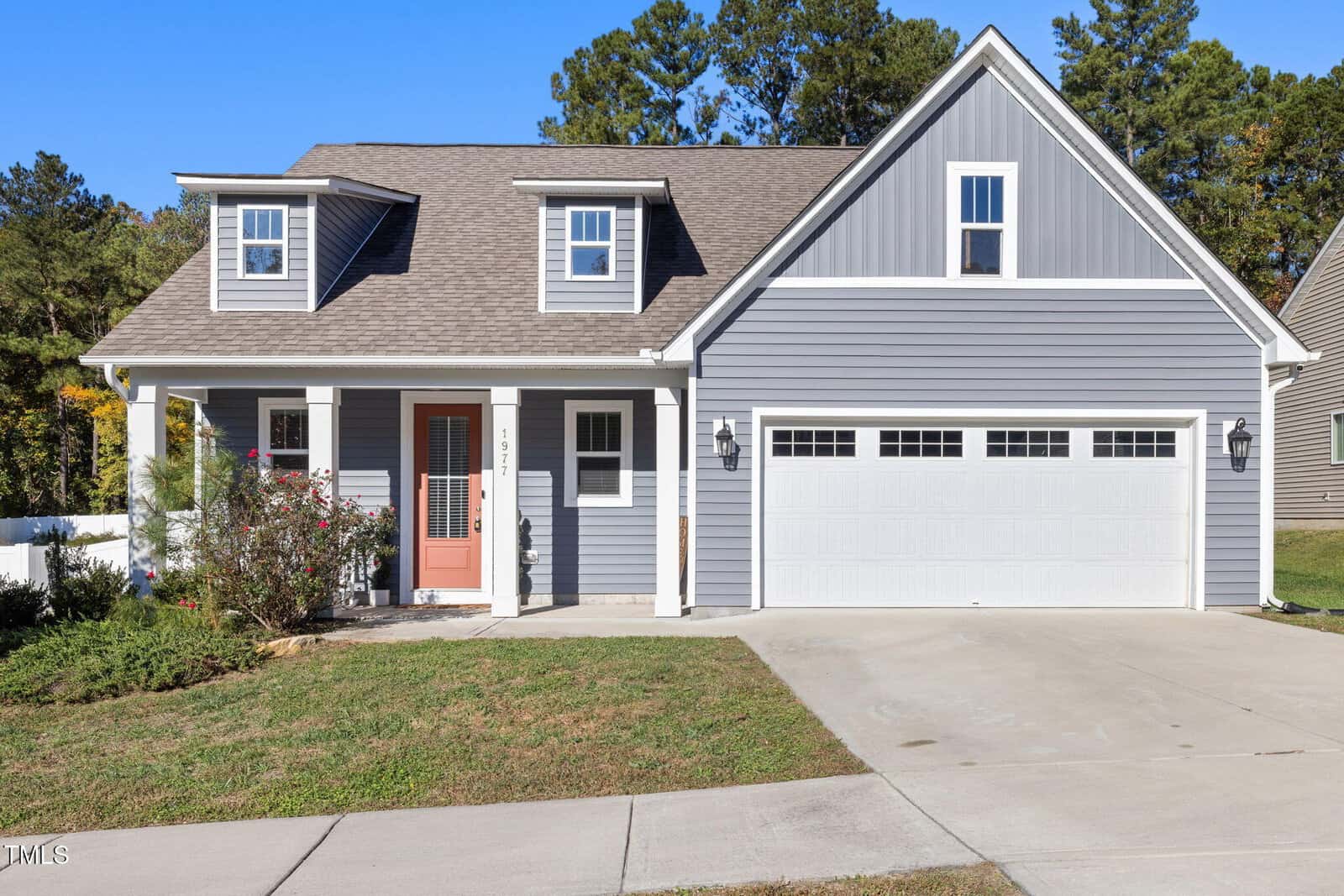 Located at 1977 Sugar Hill Drive in Creedmoor, this modern gray two-story house boasts a gable roof and white trim. It features a two-car garage, a charming front porch with white columns, and a coral front door. The landscaped yard with lush grass and shrubs is framed by a picturesque line of trees.