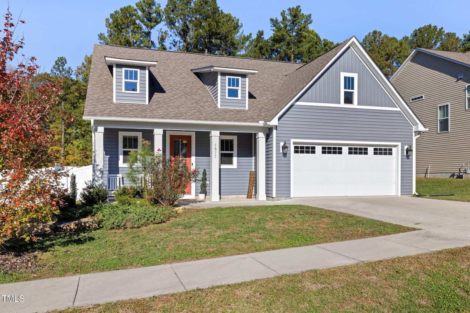 A modern, gray two-story house with white trim and a sloped roof. It features a front porch with columns and a two-car garage. A well-maintained lawn and a few small bushes decorate the front yard. Trees and another house are visible in the background.