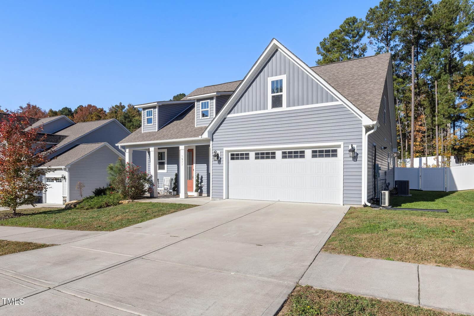 A modern, two-story gray house with white trim. It features a large garage door, a red front door, and a covered porch. The driveway is spacious, and the yard is well-maintained with some trees in the background. The sky is clear and blue.