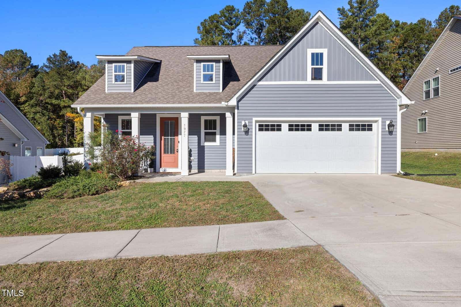 A modern, two-story gray house with white trim, featuring a spacious garage and a red front door. The home sits on a manicured lawn and has a paved driveway. The background is filled with green trees under a clear blue sky.