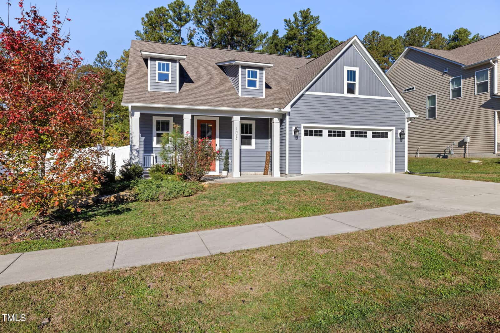 A modern suburban house with gray siding, a dormer window, and a two-car garage. The front yard has a concrete driveway and a small garden with a tree and shrubs. The house is set against a backdrop of green trees under a clear blue sky.