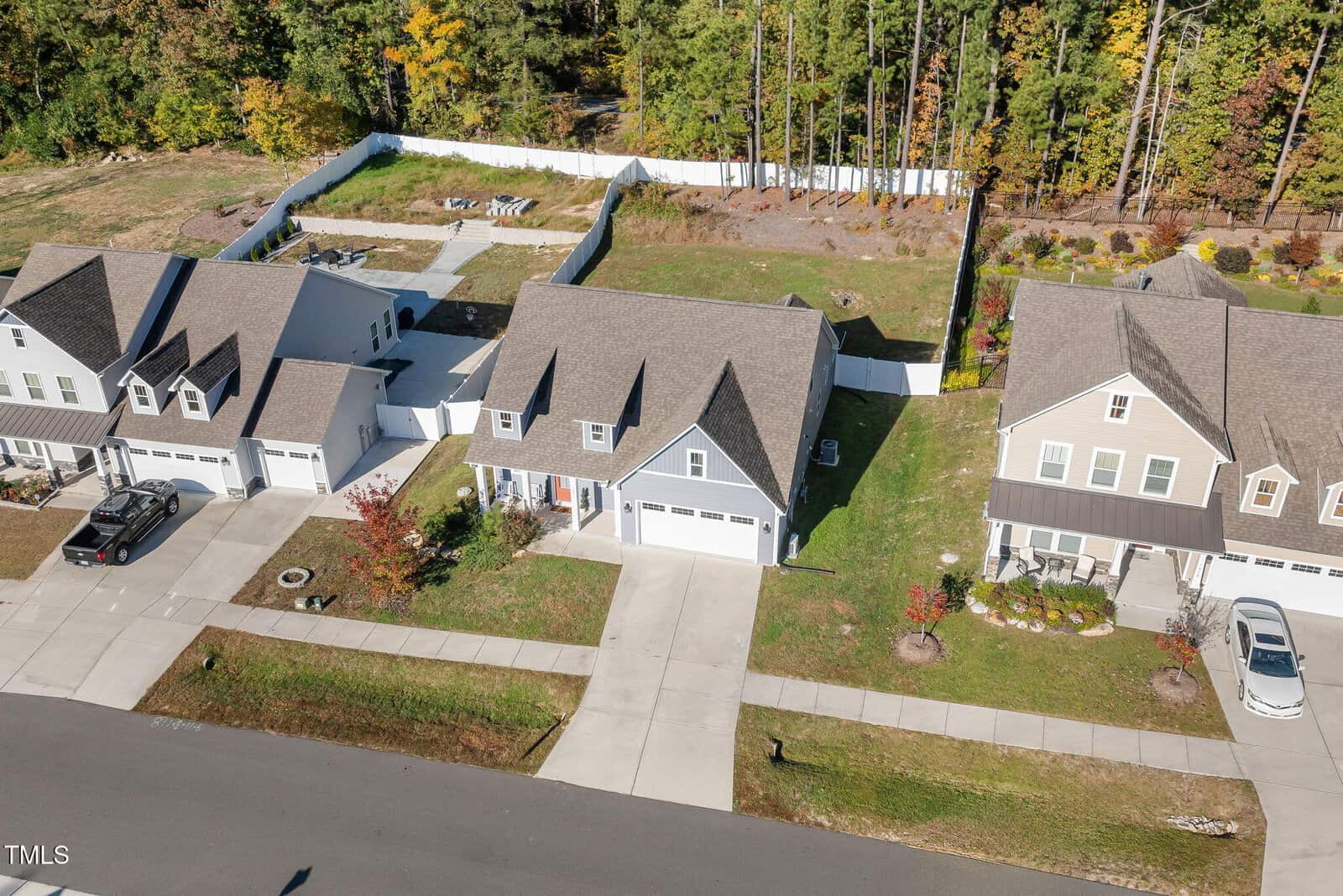Aerial view of a suburban neighborhood with three houses. Each house has a driveway and a garage. The area is bordered by trees and has a fenced backyard. The road runs horizontally in the foreground.