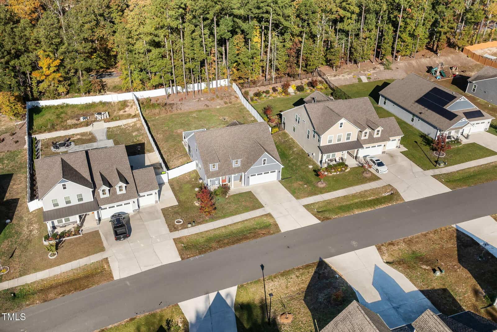 Aerial view of a suburban neighborhood showing four houses along a street. Each house has a driveway, garage, and front yard. The backyards are visible, bordered by a white fence and a row of trees. The setting is lush and green.