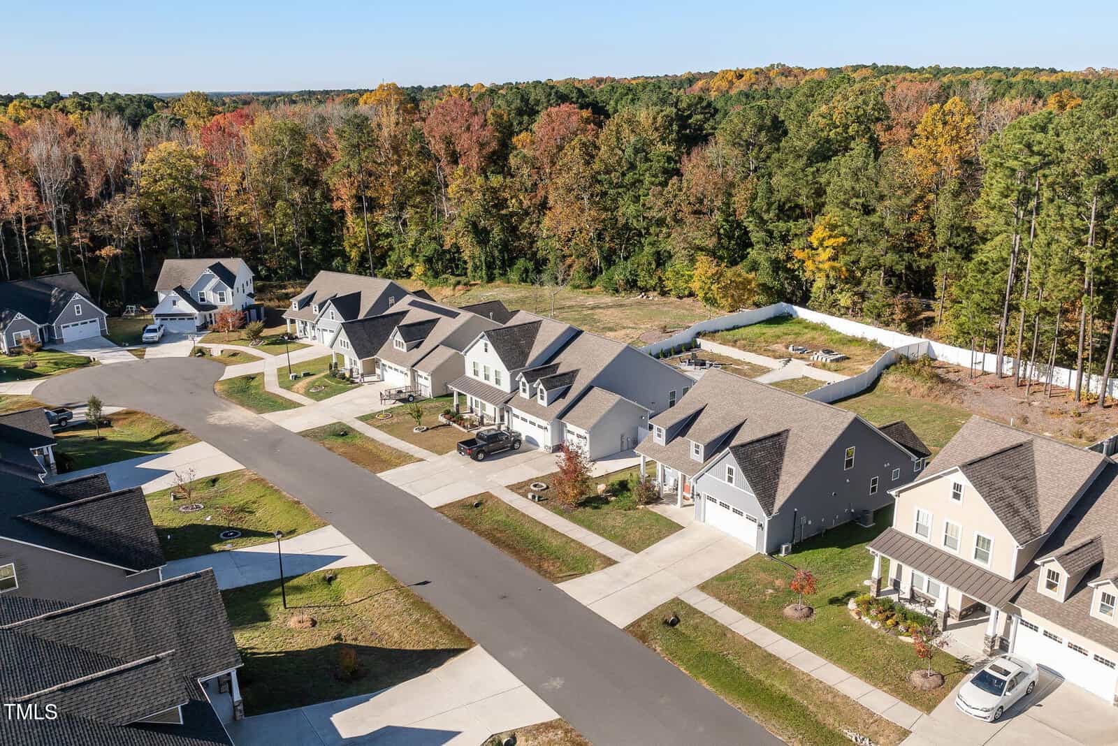 Aerial view of a suburban neighborhood with several houses along a curved street. Each house has a driveway and front yard. The area is surrounded by a forest with trees in autumn colors. A white fence partially encloses an undeveloped plot.
