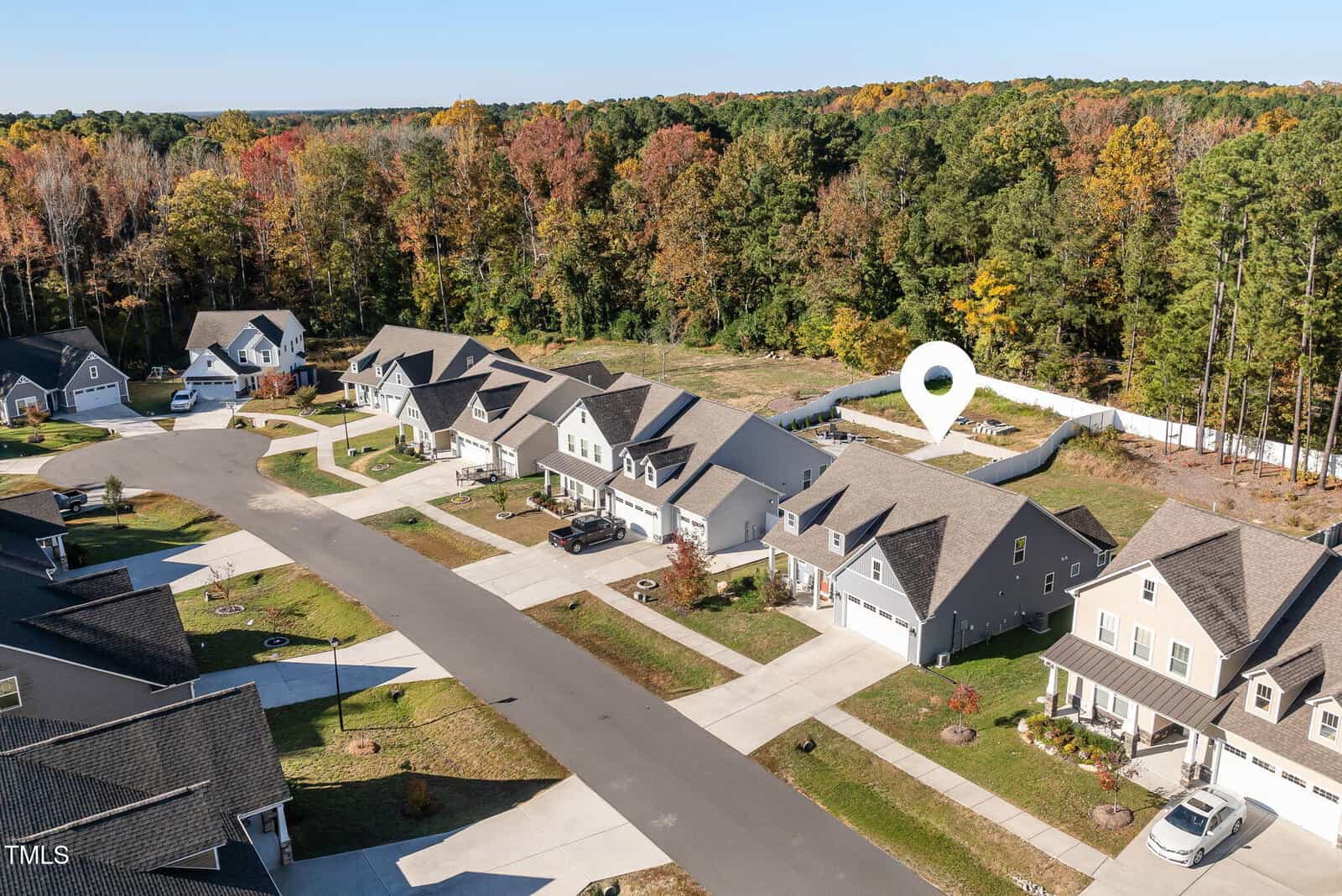 A suburban neighborhood with modern houses lined up along a curved street. There is a small wooded area with autumn-colored trees in the background. A large white location marker is positioned over one of the houses.
