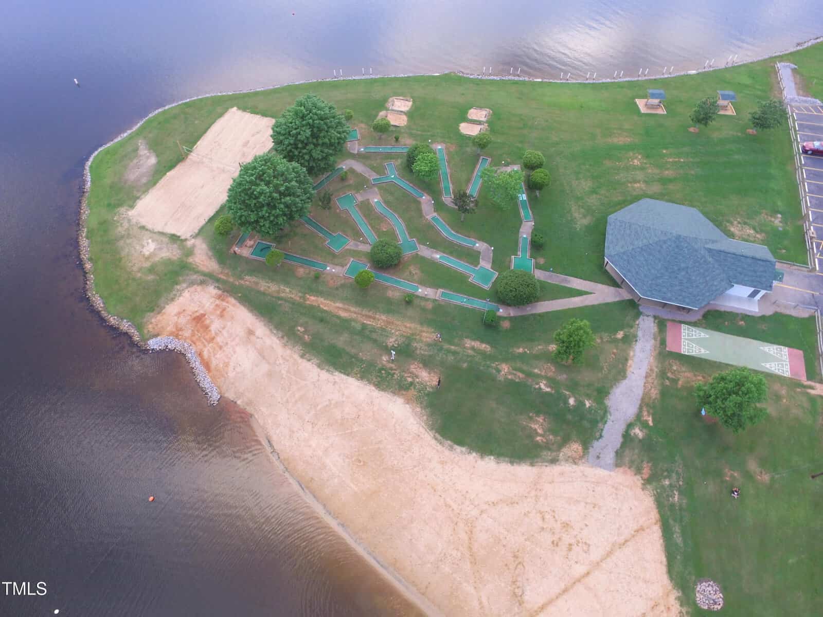 Aerial view of a mini golf course with green turf, surrounded by trees and grassy areas, next to a sandy shoreline and a body of water. A large building and parking lot are visible to the right.