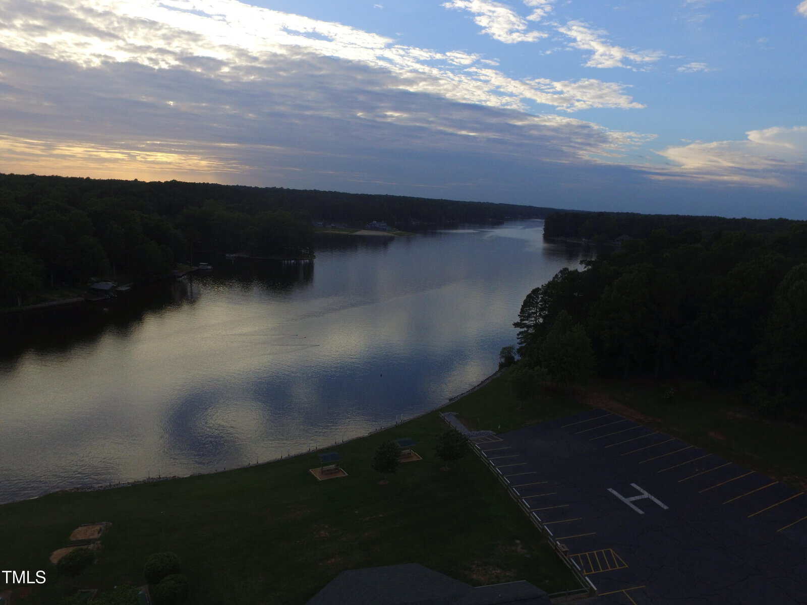 A calm lake at sunset with trees lining the shore, an empty parking lot with an “H” for helicopter landing, and partly cloudy skies reflecting on the water.