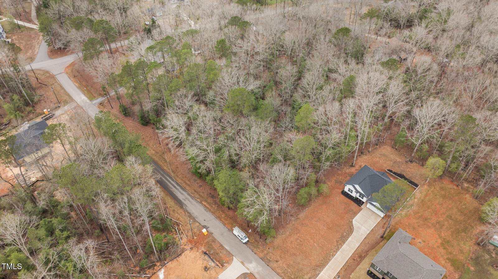 Aerial view of a wooded residential area with a road running through the center, one house with a driveway is visible on the right, and trees cover most of the scene.