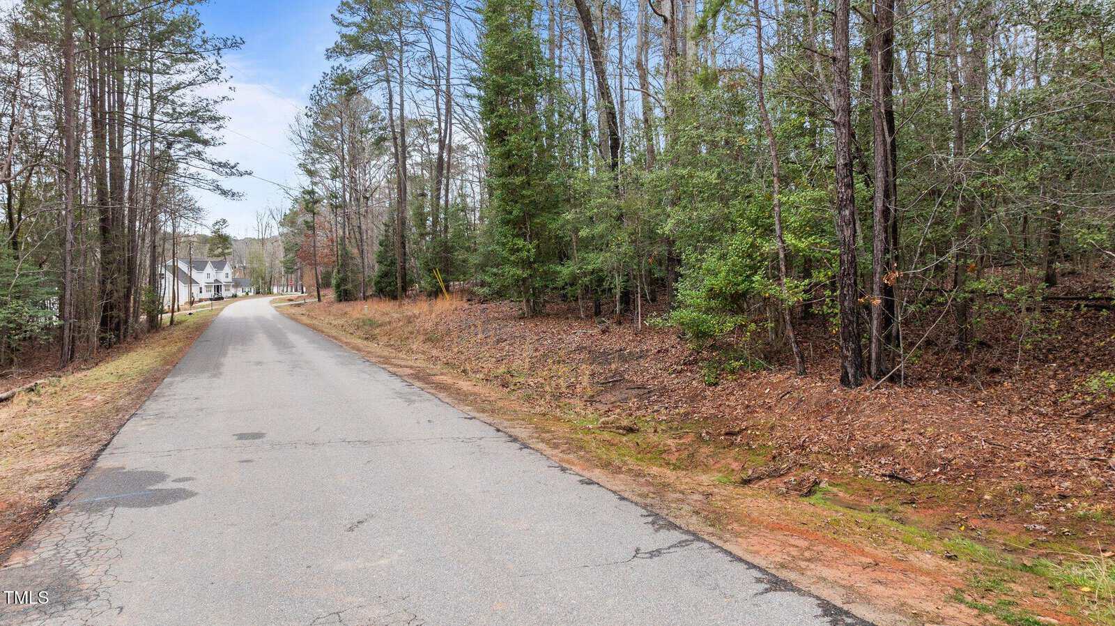 A paved road runs through a wooded area with tall trees on both sides; a white house is visible in the distance at the end of the road under a partly cloudy sky.
