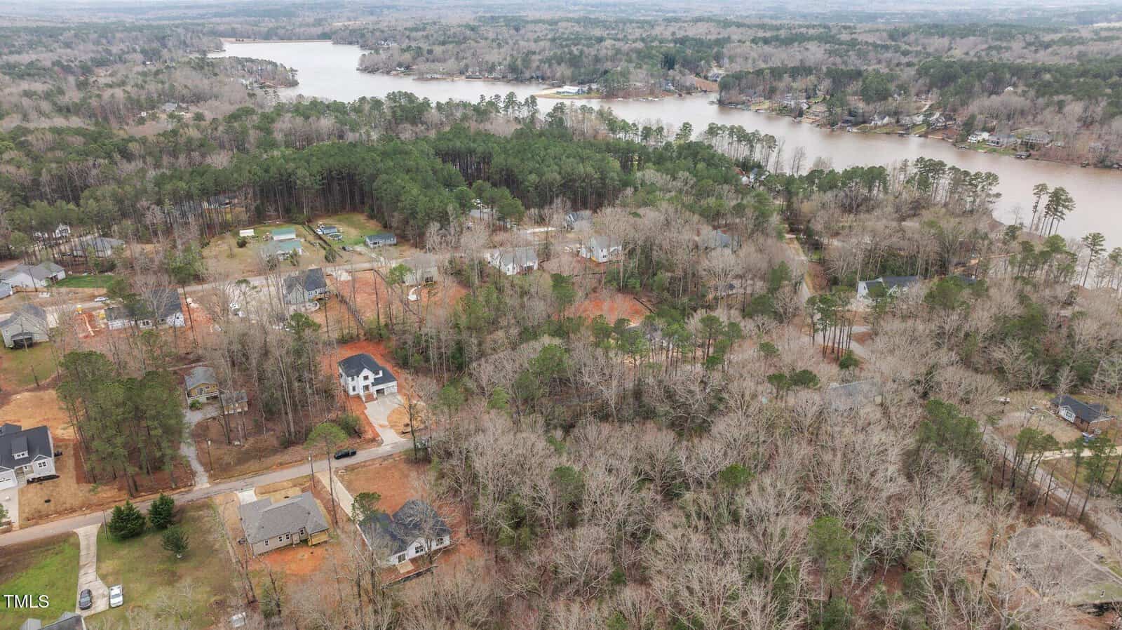 Aerial view of a residential neighborhood surrounded by dense trees, with houses spread out along winding roads and a large lake visible in the background. The area appears to be semi-rural and wooded.