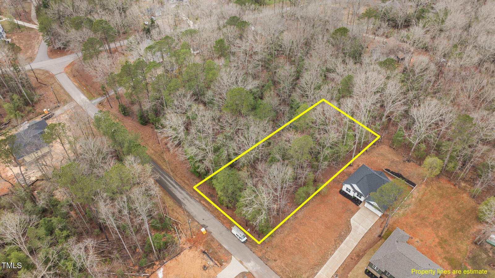A rectangular wooded lot outlined in yellow is situated between a paved road and a house with a driveway. Surrounding areas include more trees, roads, and nearby houses. Text notes property lines are an estimate.