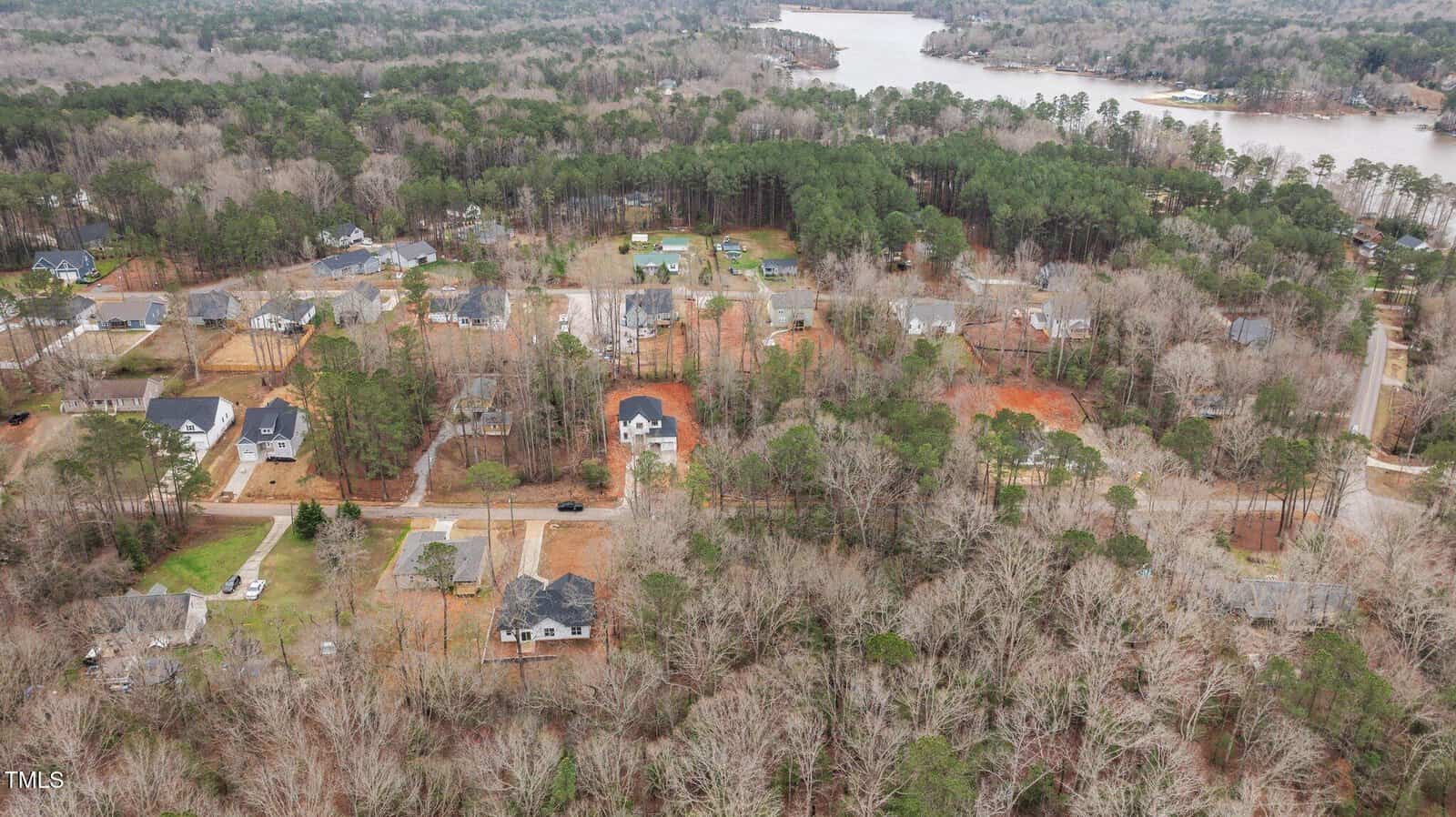 Aerial view of a residential neighborhood with scattered houses surrounded by trees; a lake is visible in the background, and some areas show patches of red soil and pale grass.