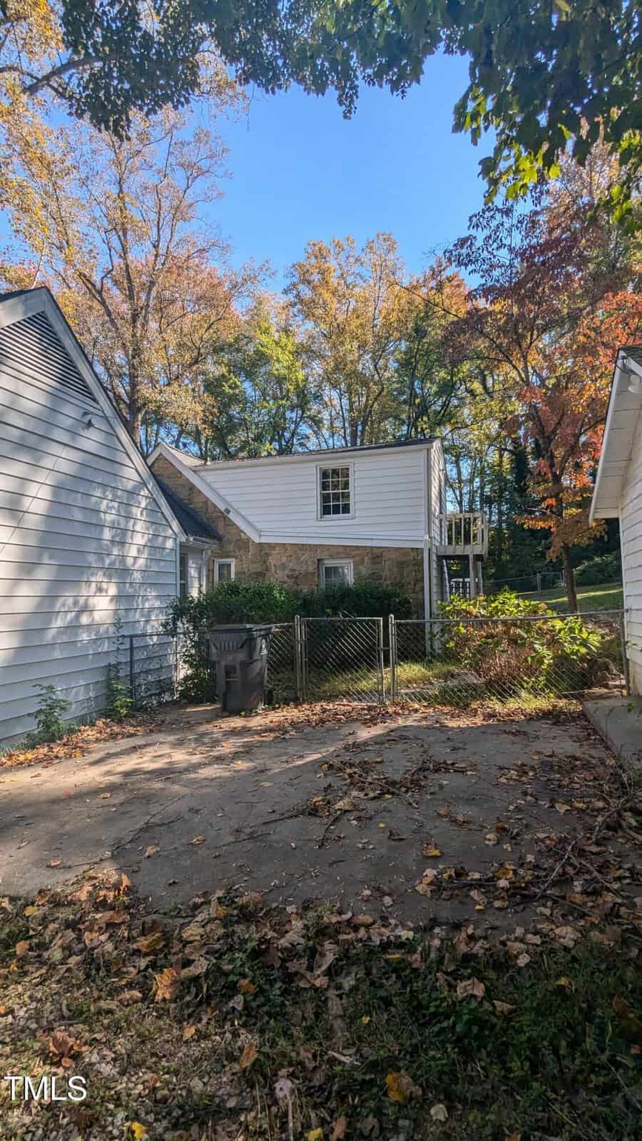 A backyard with a concrete patio surrounded by fallen leaves. There are white houses on either side with a fence in the middle, and trees with autumn foliage in the background under a clear blue sky.