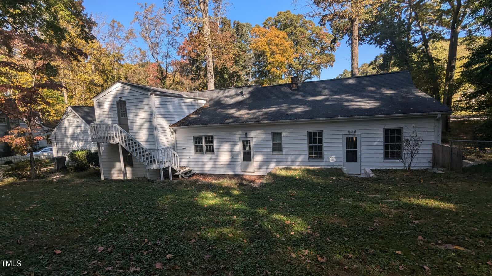 A white wooden house with a slanted roof stands surrounded by tall trees with autumn-colored leaves. There is an outdoor staircase leading to an upper entrance on the left side. The yard is grassy with some patches of shade.