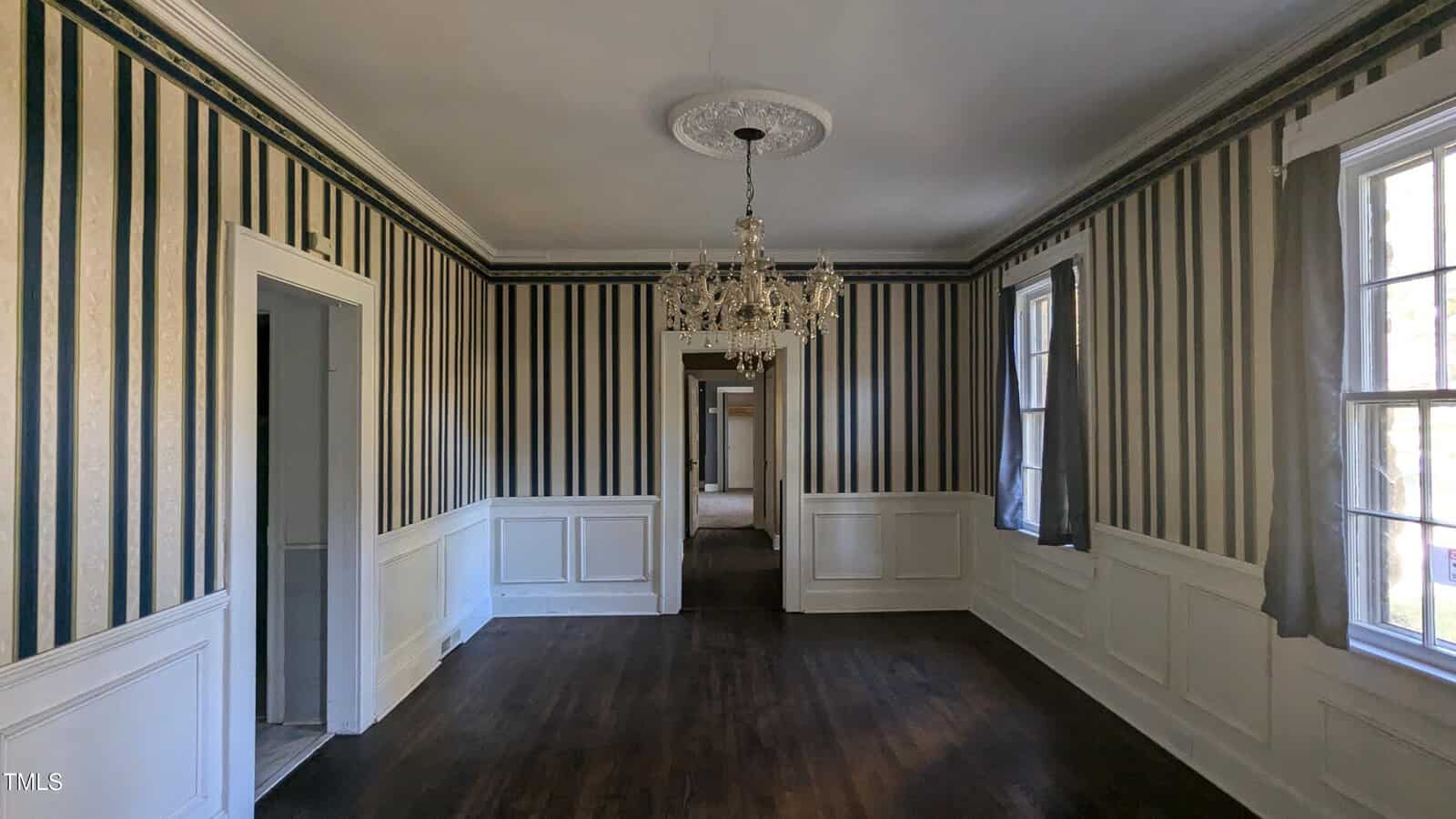 A dimly lit room with dark wooden floors and striped wallpaper in black and white. A crystal chandelier hangs from the ceiling. Natural light enters through two large windows with partially drawn curtains. The room has white wainscoting and molding.