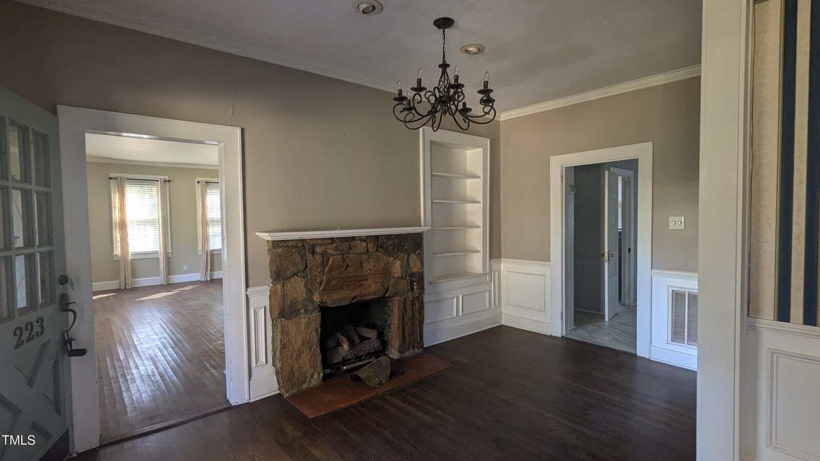 Interior of an empty room with a wooden floor, a stone fireplace, and built-in shelves. A dark chandelier hangs from the ceiling. A door on the left leads to another room with large windows. The walls are painted in neutral tones.