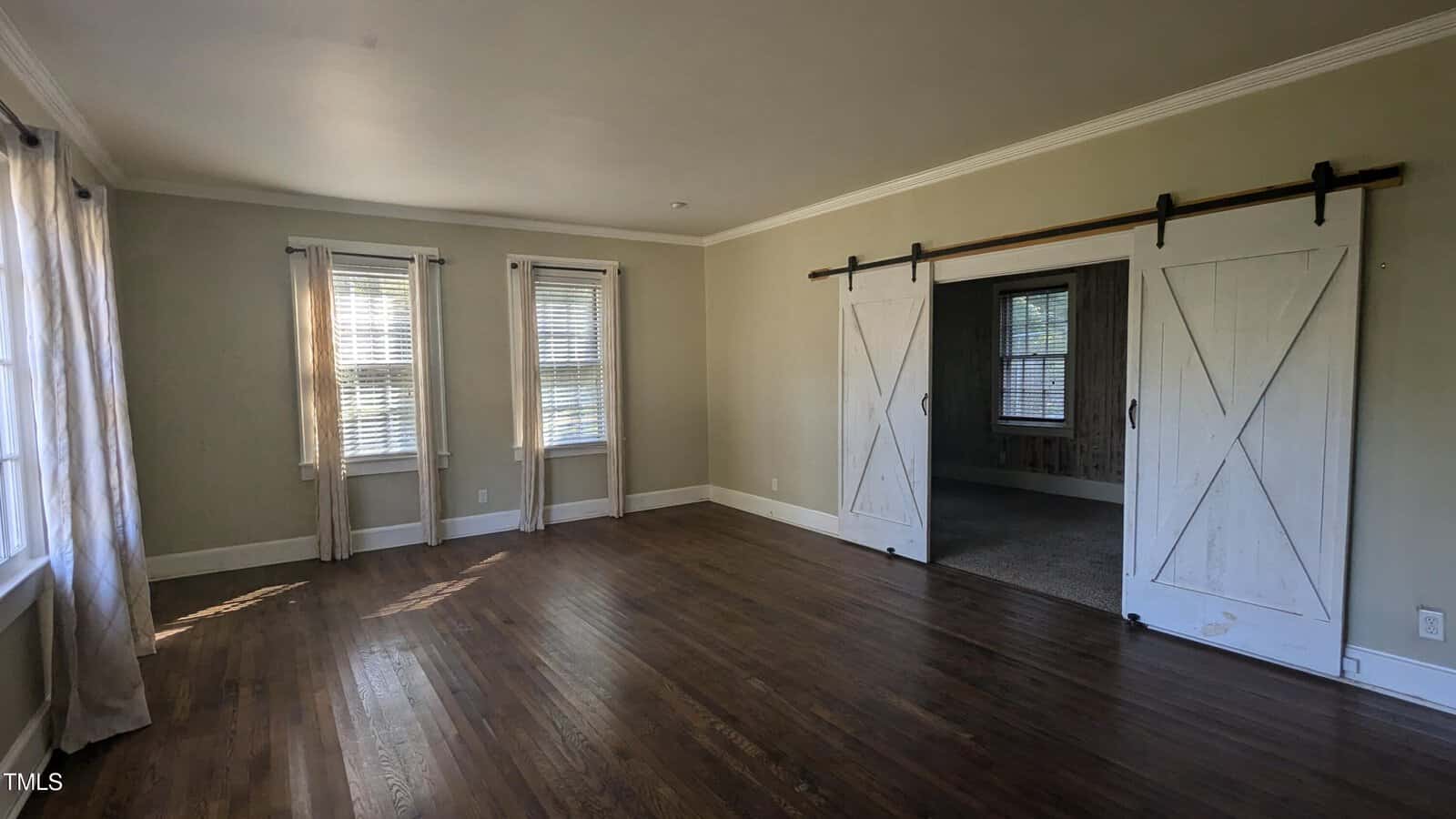 A spacious, empty room with wooden floors and light-colored walls. Large windows with white blinds allow natural light to stream in. A sliding barn door separates the room from an adjacent carpeted area.