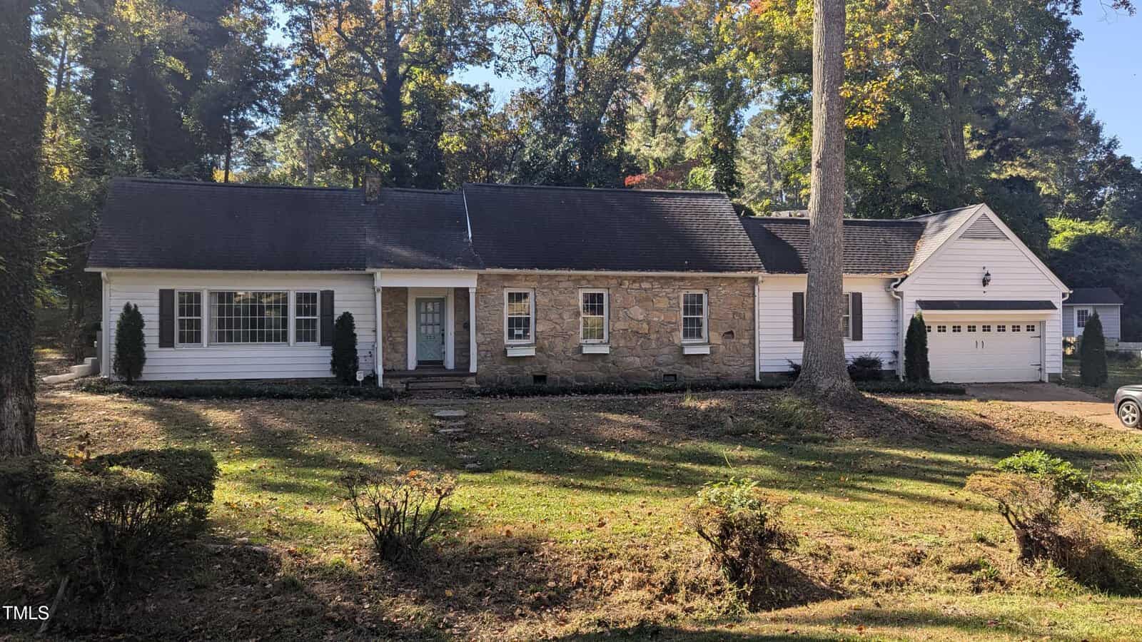 Located at 223 Willowood Drive in Henderson, this single-story house features a white exterior with stone accents. It boasts a dark roof, large front windows, and a door with sidelights. A detached garage sits to the right, surrounded by a grassy yard and tall trees.