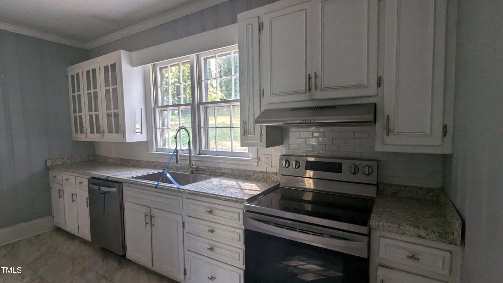 A kitchen with white cabinets, granite countertops, and a stainless steel oven. There is a dishwasher next to the sink under a window with a view of greenery outside. The backsplash features white subway tiles.