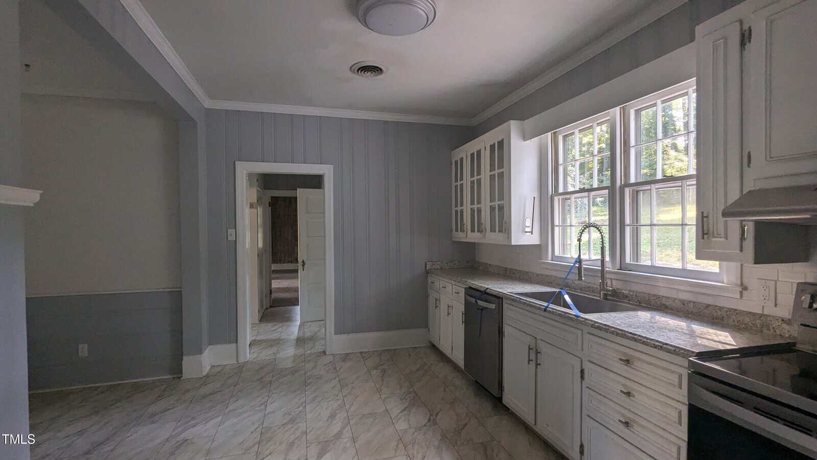 A kitchen with white cabinets and drawers, gray walls, and marble-patterned flooring. It features large windows, a stainless steel dishwasher, and an oven. Light streams through the windows, illuminating the space.