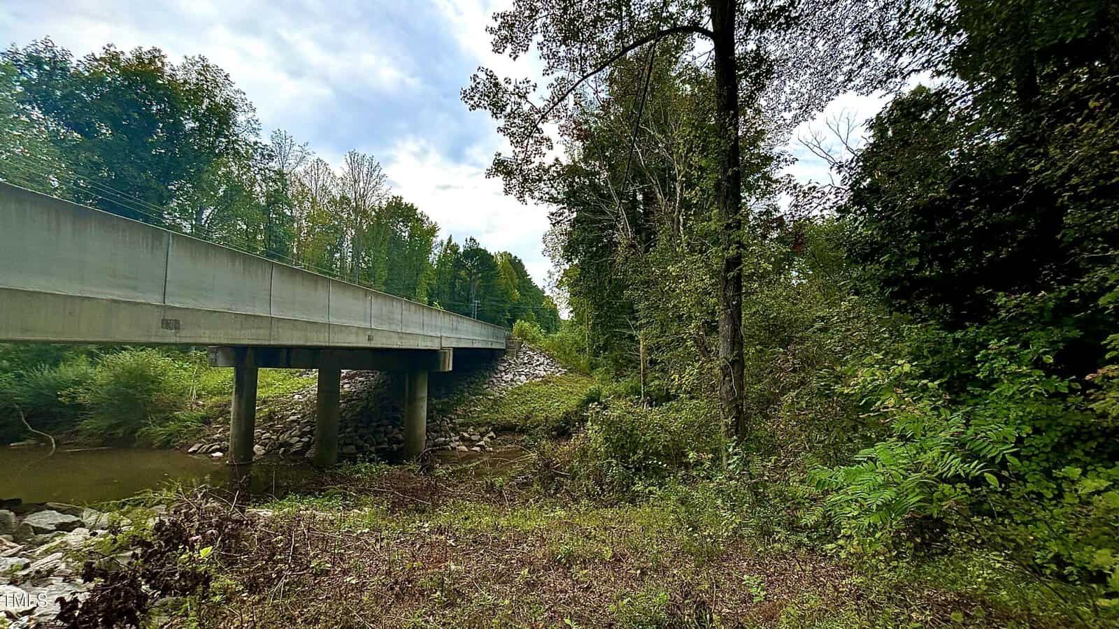A concrete bridge stretches over a narrow creek, surrounded by dense, green foliage and tall trees. The foreground shows a grassy, slightly overgrown area leading to the bridge. The sky is cloudy, adding a serene atmosphere to the scene.